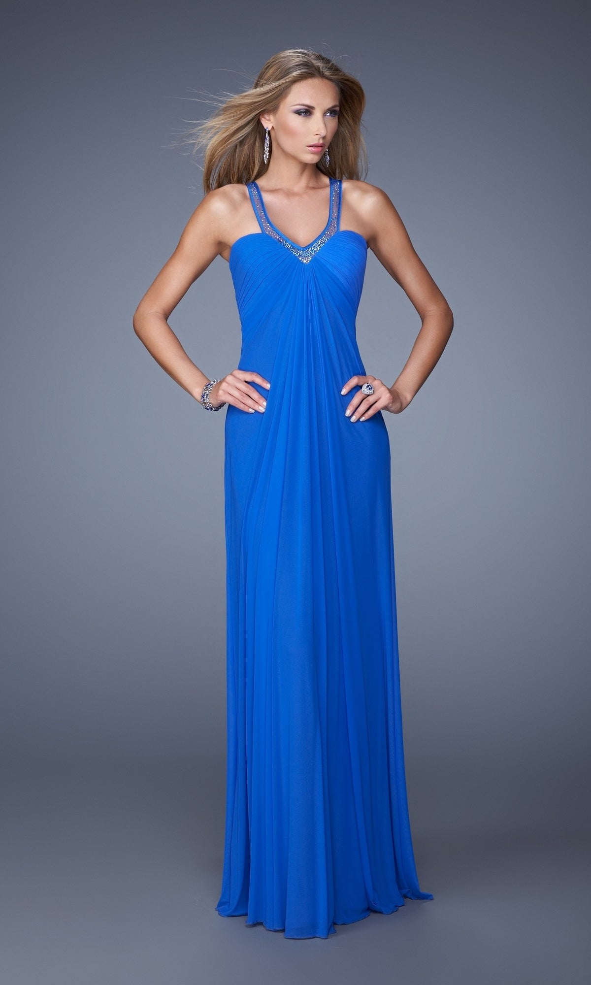 A woman poses against a gray background, hands on hips, wearing the La Femme Dress 20903—a floor-length royal blue gown with embellished straps.