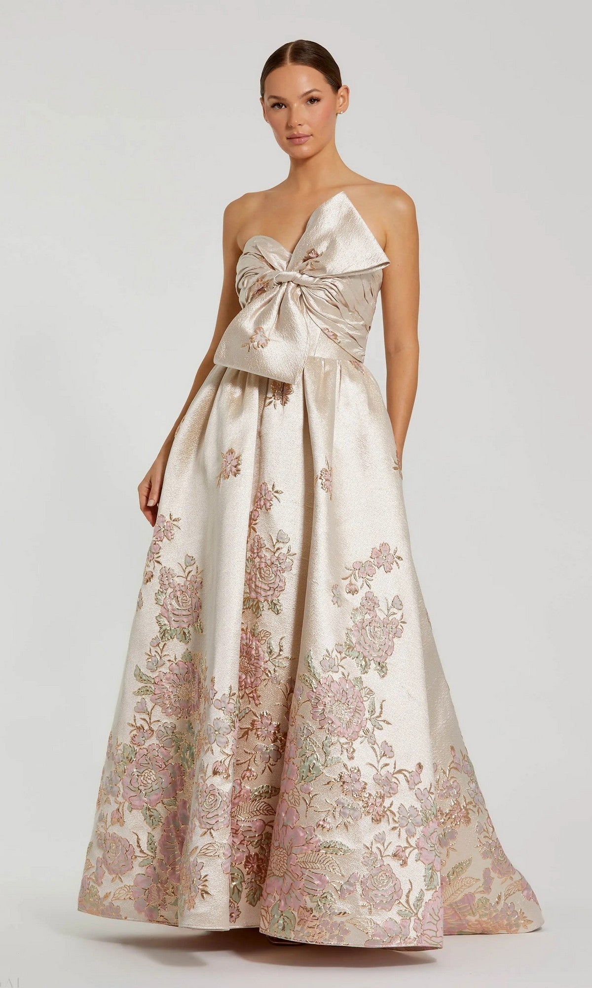 A woman stands against a plain background in the Mac Duggal 20974 Long Formal Dress in Champagne Multi, featuring a floor-length silhouette, large bow bodice, and floral embroidery.