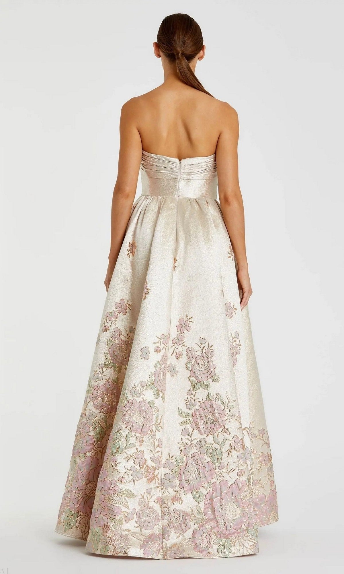 A woman with straight brown hair in a ponytail stands elegantly with her back to the camera, wearing the Mac Duggal 20974—a strapless, floor-length cream floral ballgown with pink and green patterns.