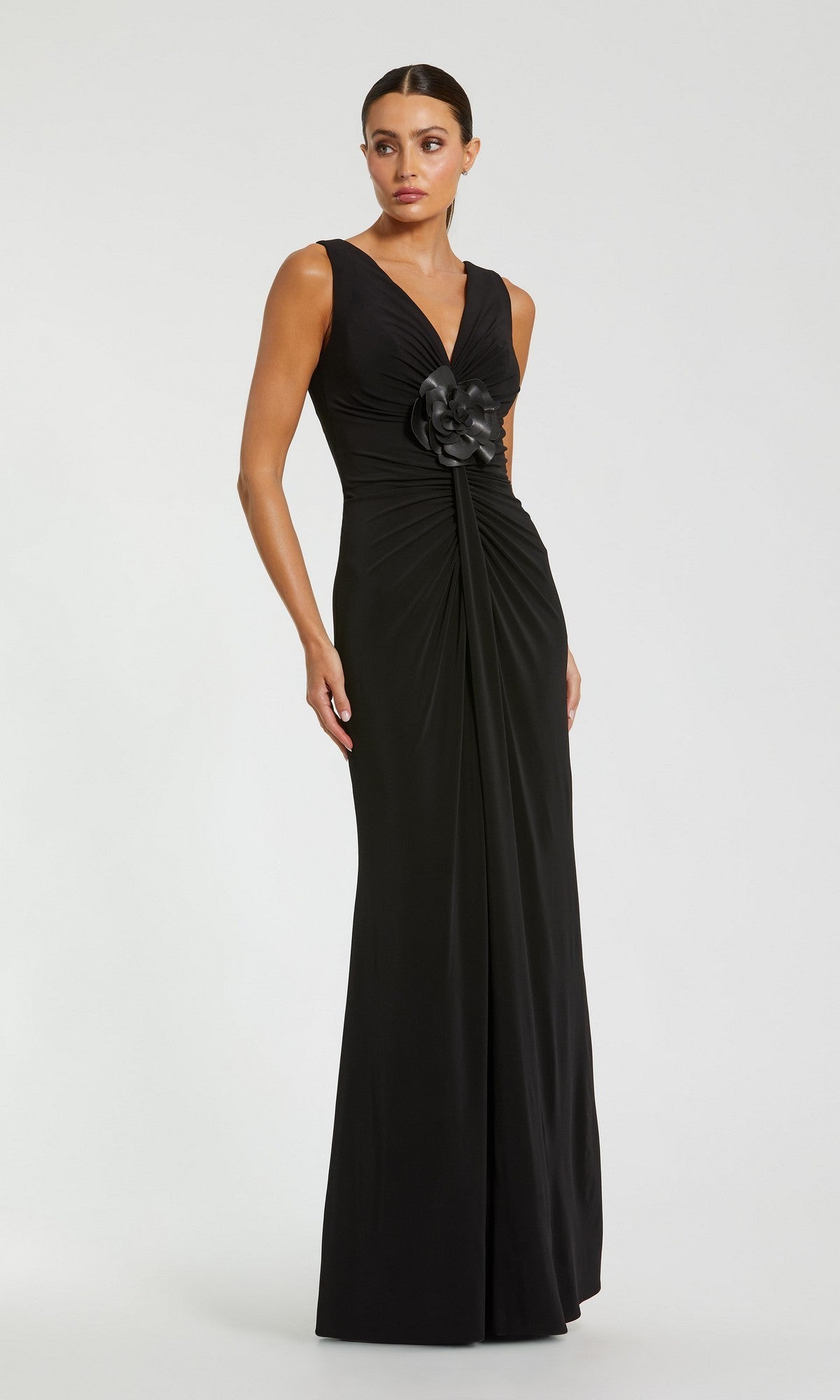 A woman stands against a plain light background wearing the Mac Duggal 21005, a sleeveless, floor-length black formal dress featuring a V-neckline, ruched detailing, and a 3D flower accent at the waist.