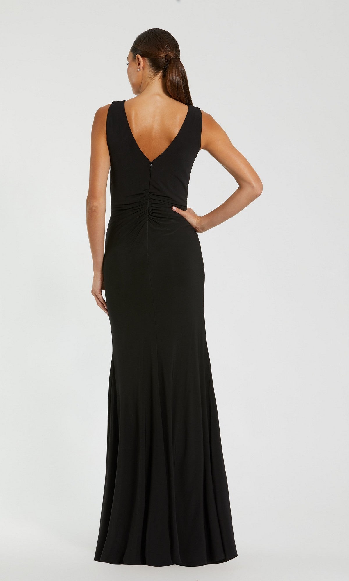 A woman wearing the Mac Duggal 21005 long formal dress—a sleeveless, floor-length black ruched gown with a deep V-back—stands against a plain light background, facing away from the camera.