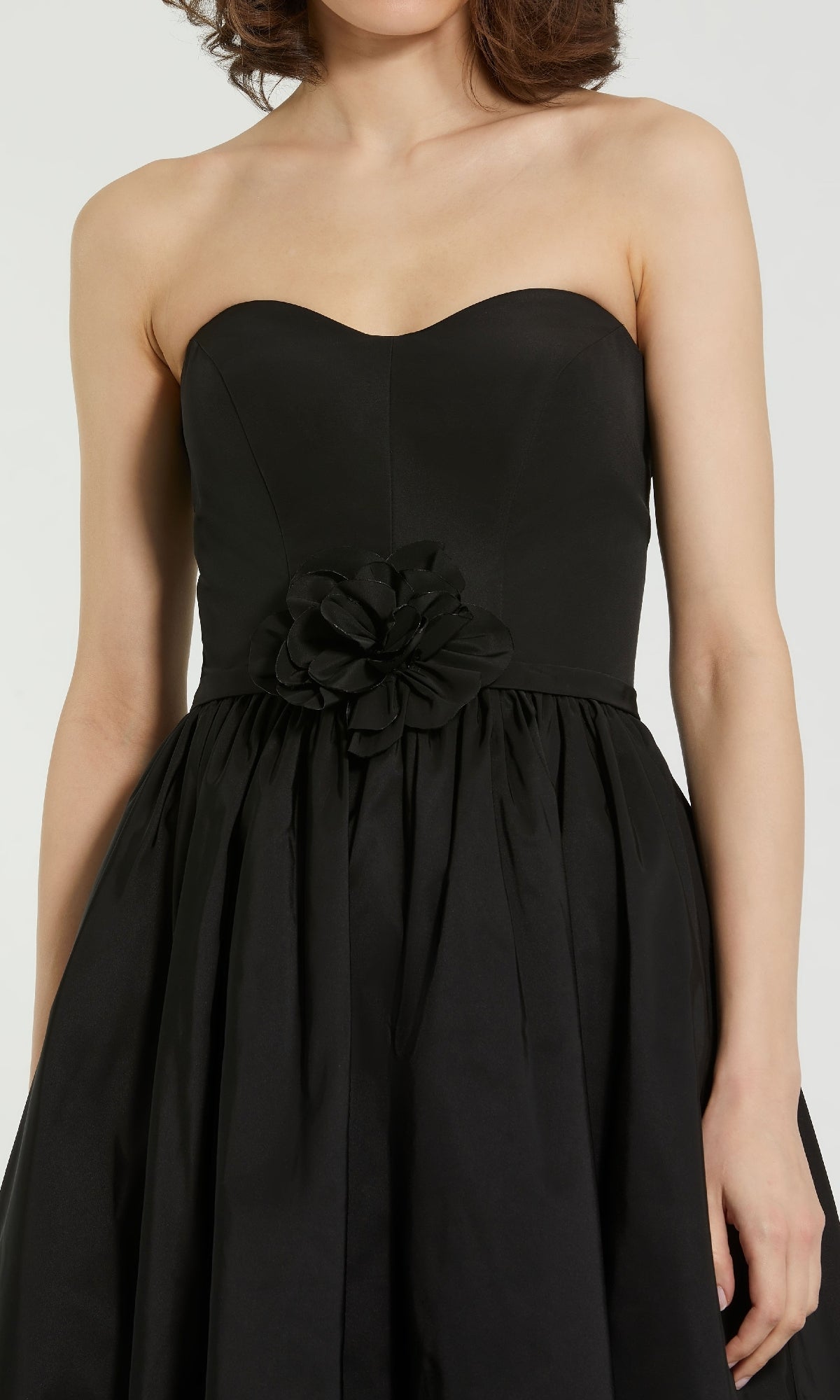 A woman wears the Mac Duggal 21023: a strapless, tea-length black midi dress with a sweetheart neckline and a bold fabric flower at the waist—an elegant semi-formal choice.