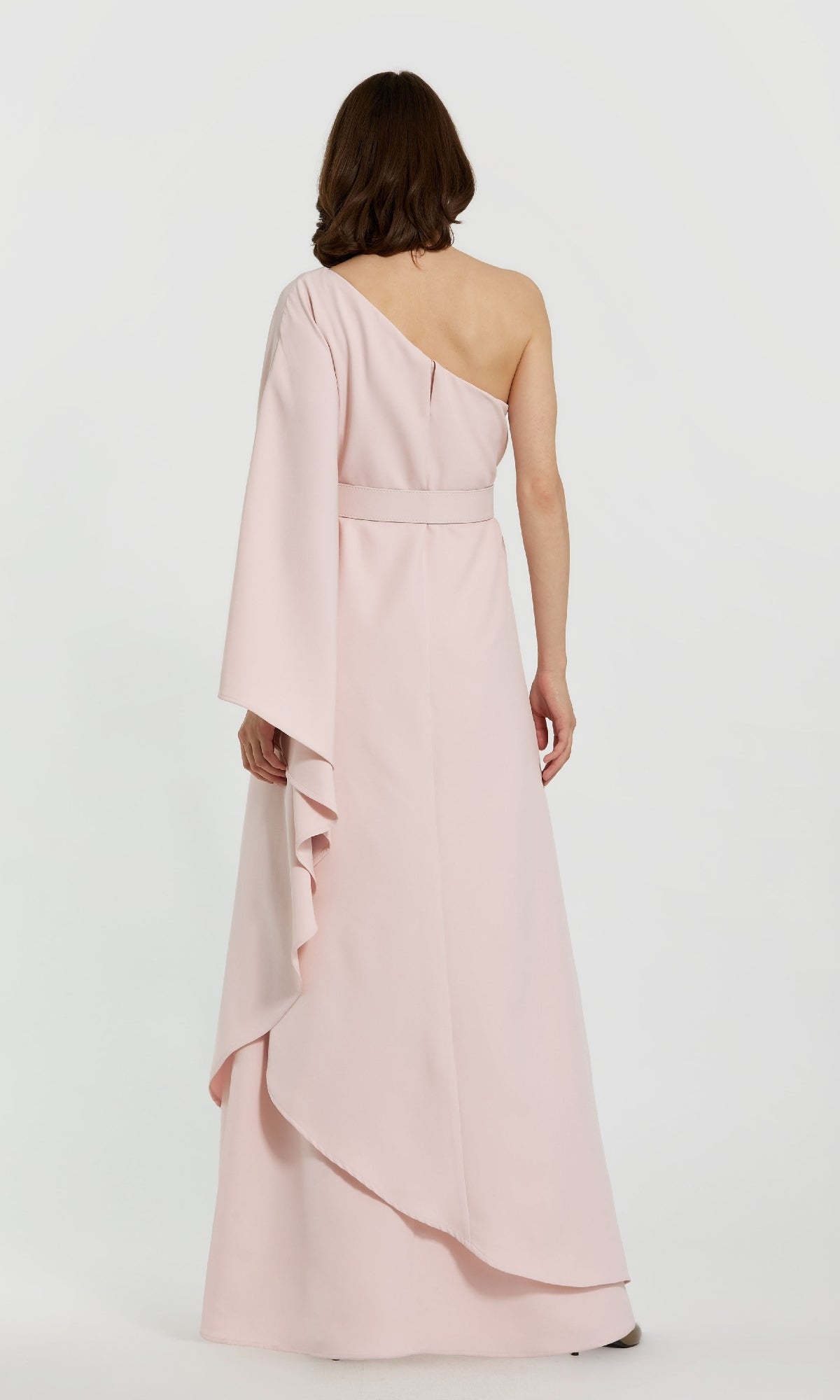 A woman stands facing away, wearing the Mac Duggal 21049—an elegant, pale pink, one-sleeve formal dress that’s perfect for the mother of the bride or any special occasion.