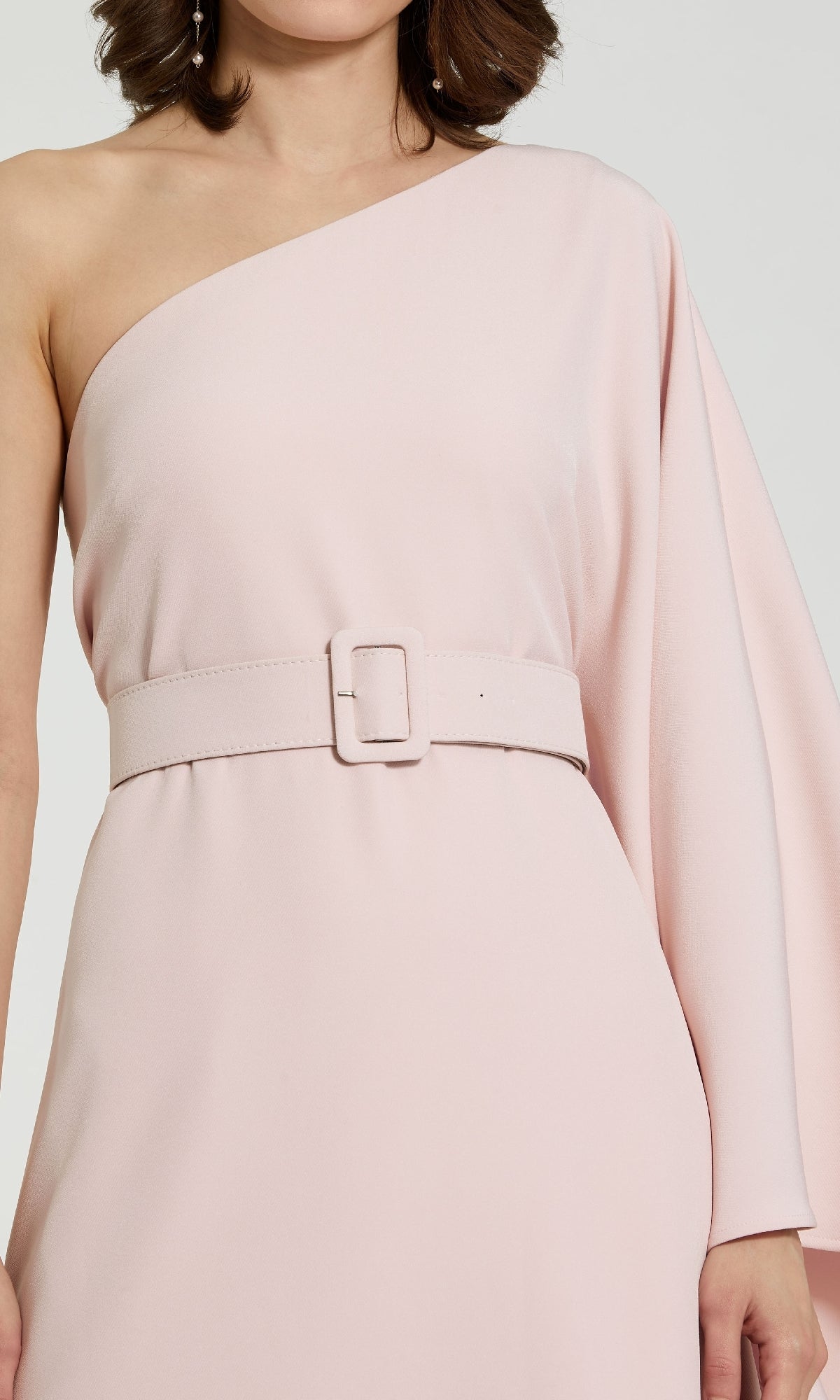 A woman models the Mac Duggal 21049 One-Sleeve Pink Formal Dress, featuring a light pink hue, matching belt, and asymmetrical sleeve—an elegant mother-of-the-bride choice. Shown from shoulders to hips.
