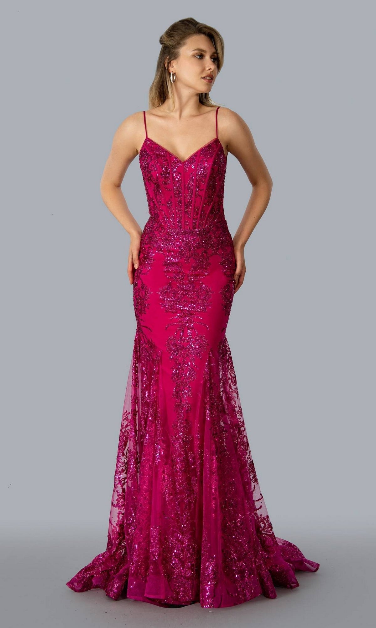 A woman stands against a plain gray background wearing the Corset-Bodice Sparkly Long Mermaid Prom Dress 21066, a fitted magenta sequin gown with lace detailing, thin straps, sheer overlay, and an embellished corset silhouette.