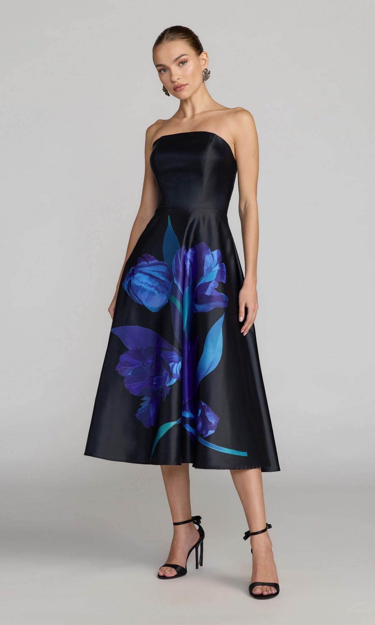 A woman is wearing the Mac Duggal 21075 Midi Floral Semi-Formal Dress in Black Multi, featuring a strapless design with large blue and purple floral prints, paired with black high-heeled sandals and earrings, standing against a plain light background. This designer dress is perfect for any floral party occasion.