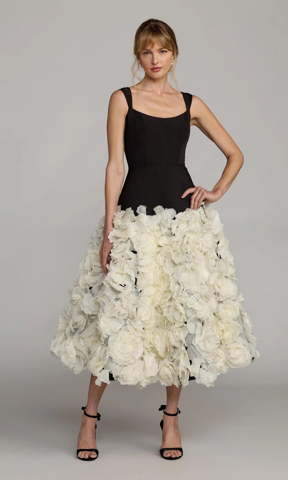 A woman wears the Mac Duggal 21080 midi wedding guest dress—black, sleeveless crepe with a voluminous skirt featuring large 3D organza flowers—paired with black high-heeled sandals.
