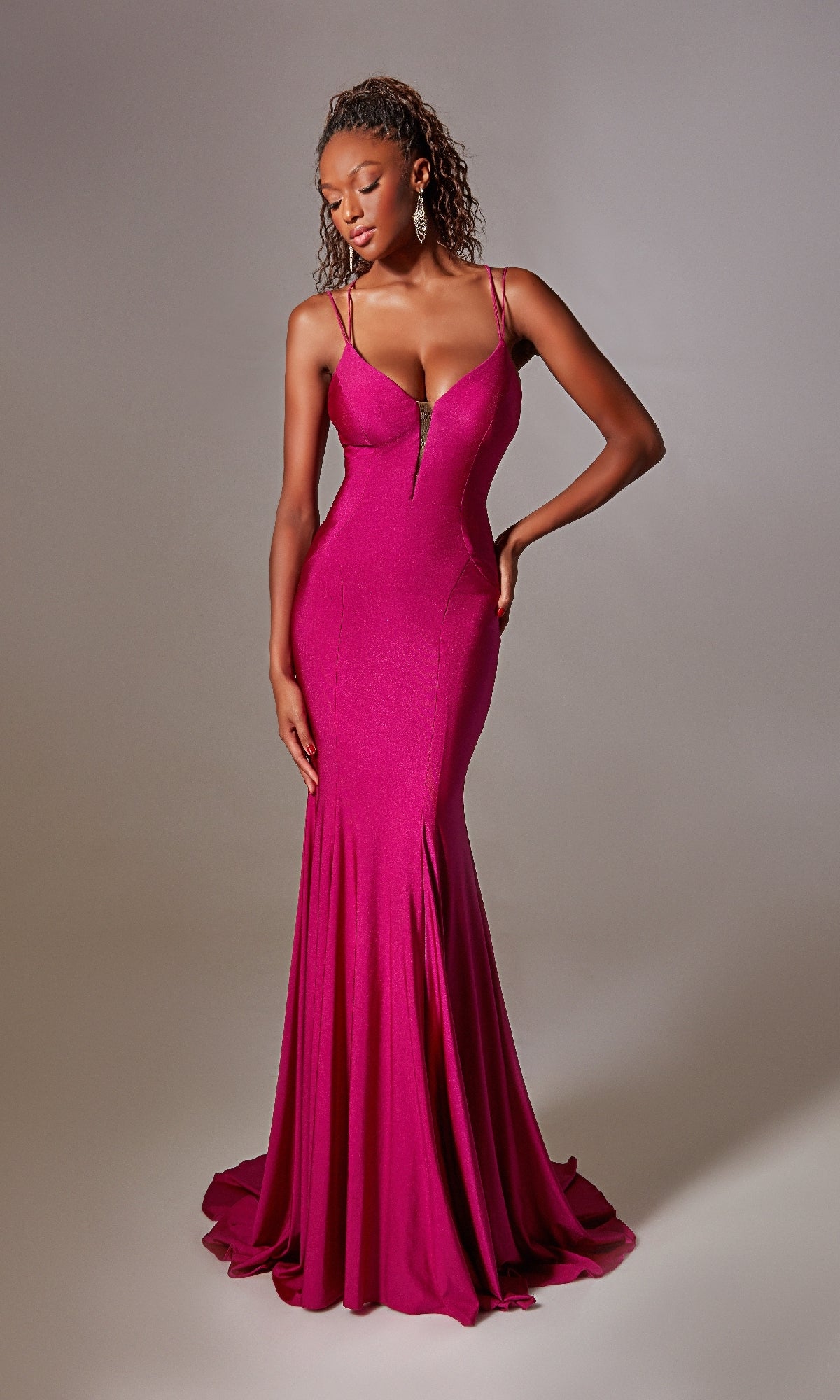 A woman poses with one hand on her hip in the Aerie Couture 210, a fitted, floor-length deep V-neck magenta prom dress featuring thin straps and a lace-up open back, standing against a plain background.