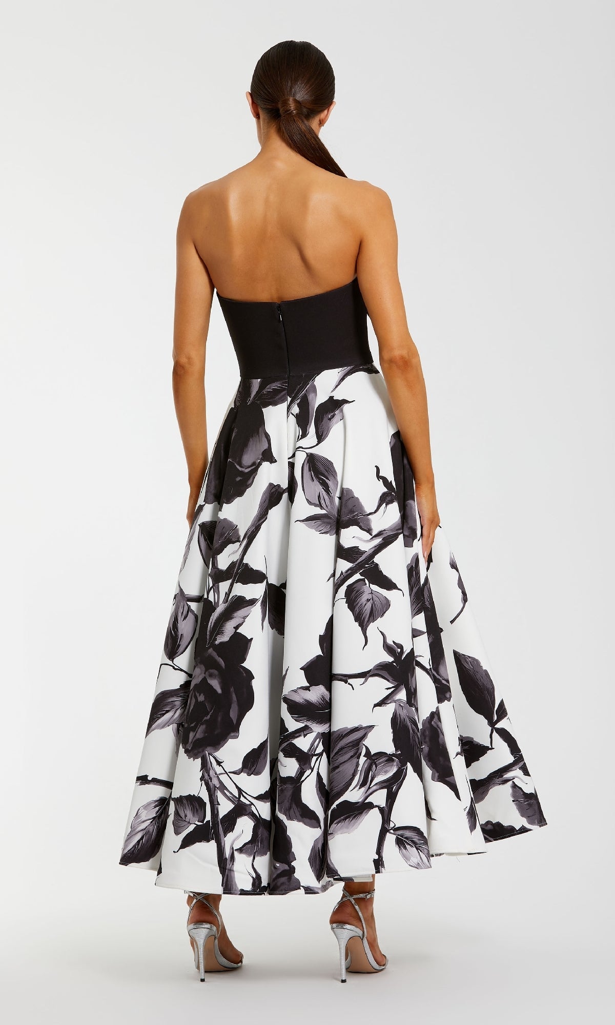 A woman stands with her back to the camera, wearing the Mac Duggal 21151 long formal dress— a strapless black and white floral evening gown—paired with silver high-heeled sandals.