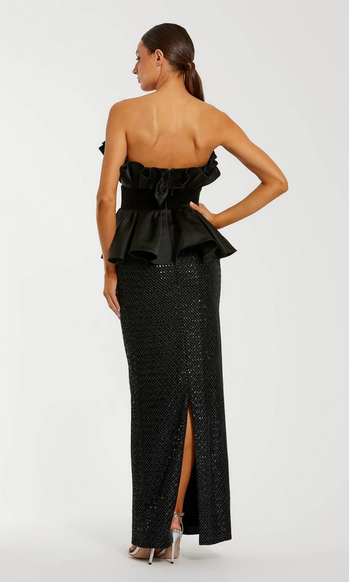 Woman faces away from the camera in the Mac Duggal 21174 long formal dress, showcasing a strapless black design with a ruffled top, sequin column skirt, back slit, and silver heels.