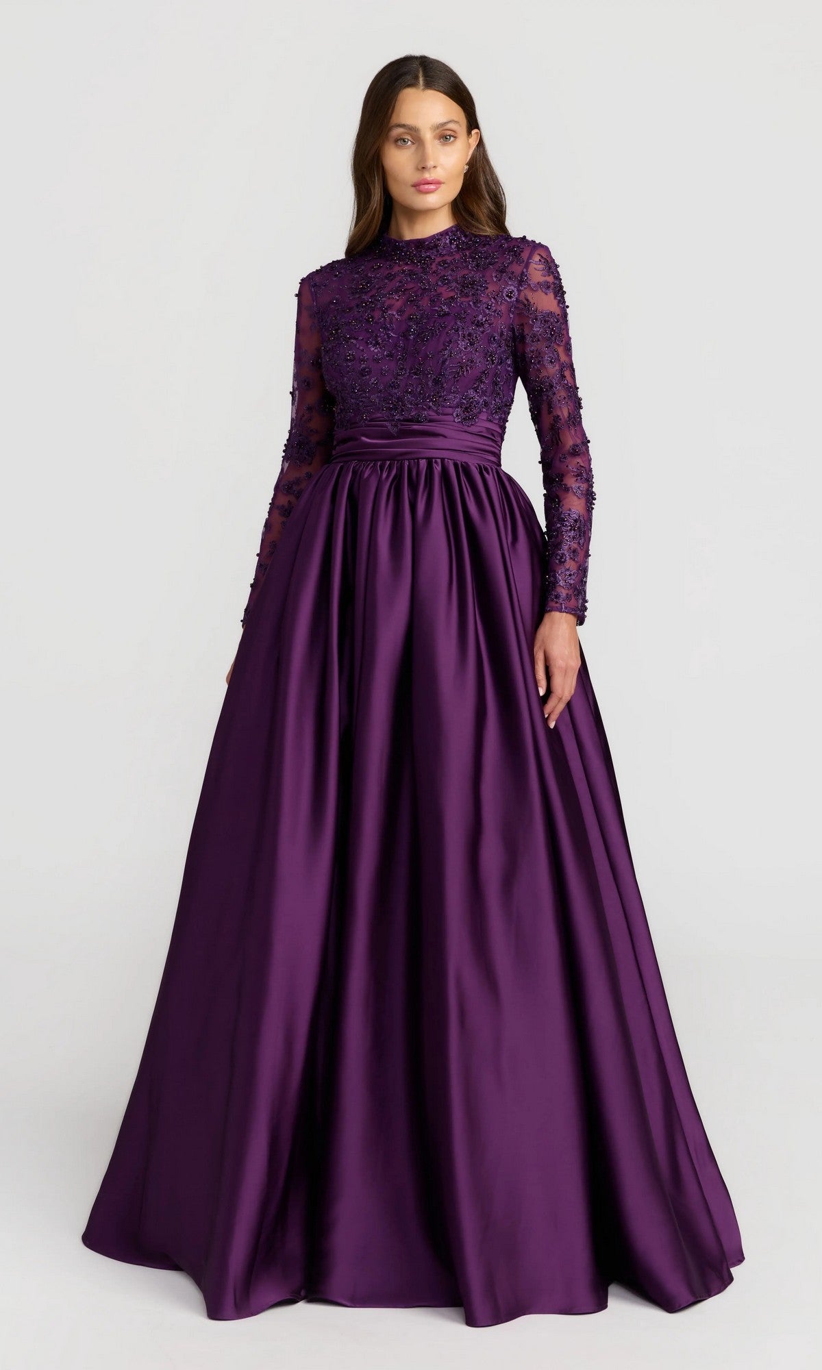 A woman wears the Mac Duggal 21189 long formal dress in aubergine, featuring long sleeves and a beaded-embroidered bodice, standing against a plain light background.