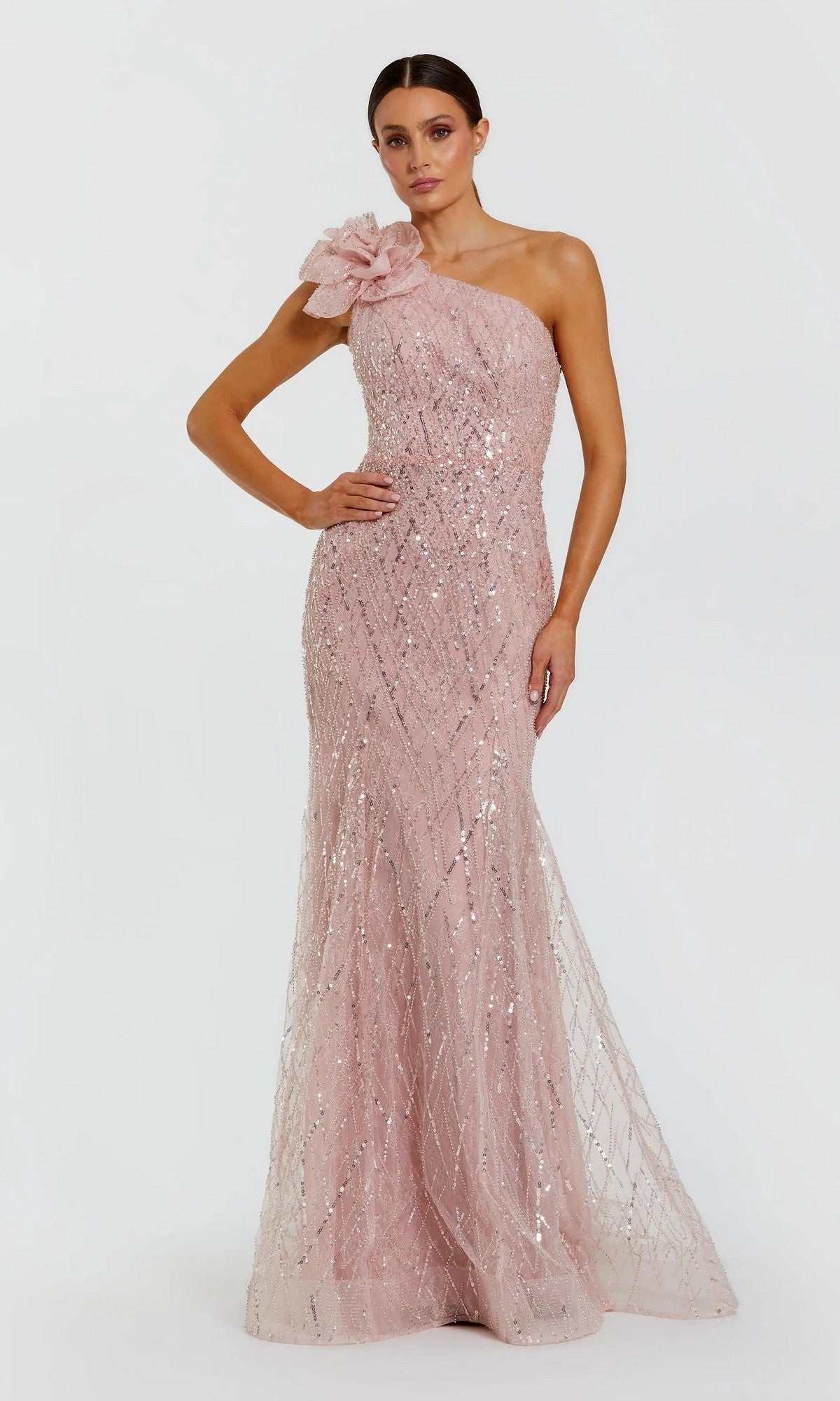 A woman wears the Mac Duggal 21199 long formal dress in rose, featuring a one-shoulder design adorned with sparkling sequins and a large floral detail on the shoulder, as she stands against a plain white background.