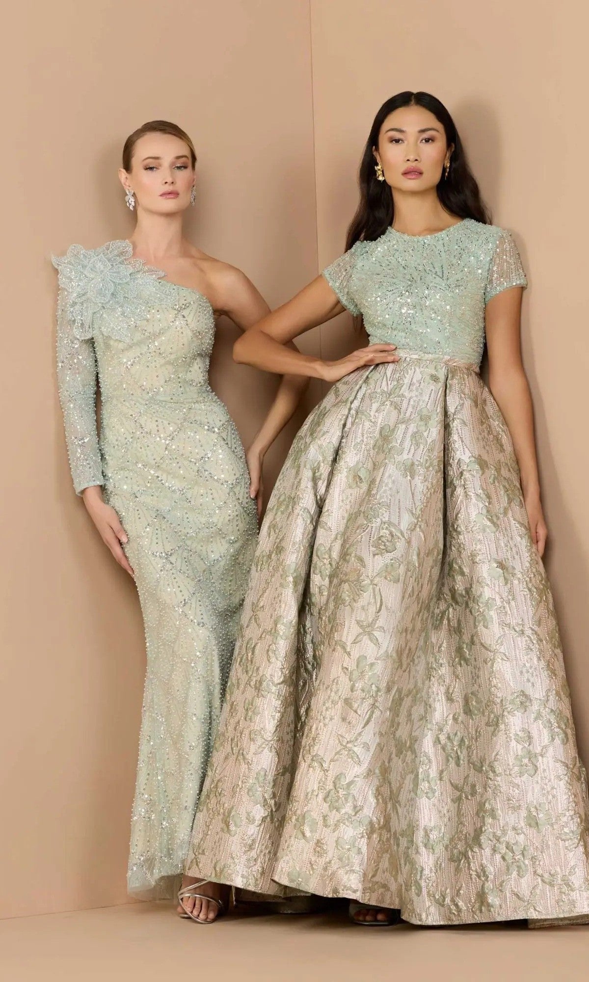 Two women pose against a beige backdrop in elegant light blue and silver gowns featuring intricate beadwork and floral designs—one dazzles in a sequin-beaded dress, while the other wears the graceful Mac Duggal 21203 long formal dress.