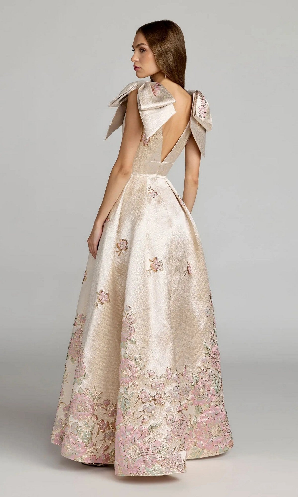 A woman wearing the Mac Duggal 21204 long formal dress—sleeveless and floor-length, featuring satin brocade fabric, a deep V-back, bow-detailed shoulders, and pink floral embroidery near the hem—stands against a plain background.