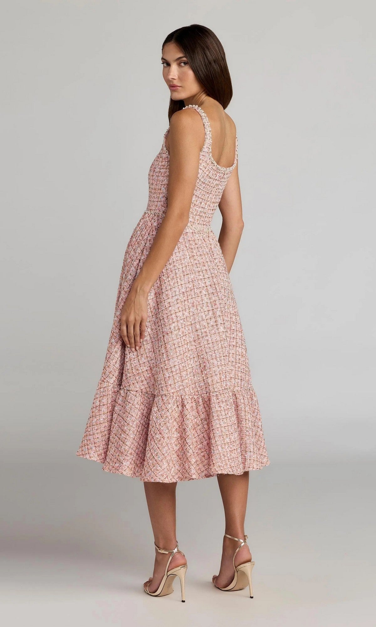 A woman stands in profile against a plain gray background, wearing the Midi Party Dress: Mac Duggal 21207—a sleeveless, pink patterned midi dress with a ruffled hem—paired with beige high-heeled sandals.