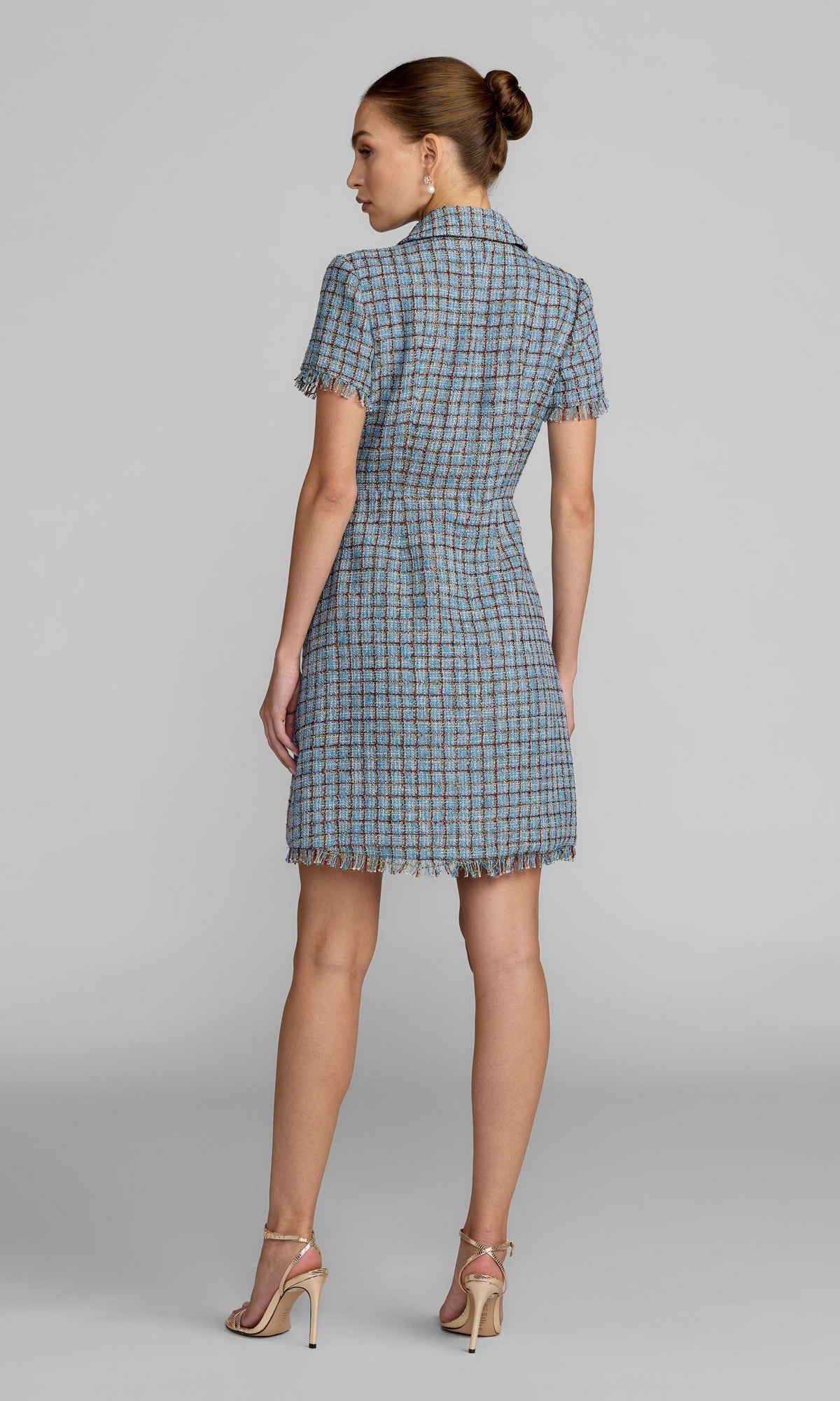 A woman stands with her back to the camera, modeling the Mac Duggal 21221 Short Boucle Day Dress—a short-sleeve blue plaid mini dress with a collared neckline—paired with beige heels and her hair in a bun, set against a plain gray background.