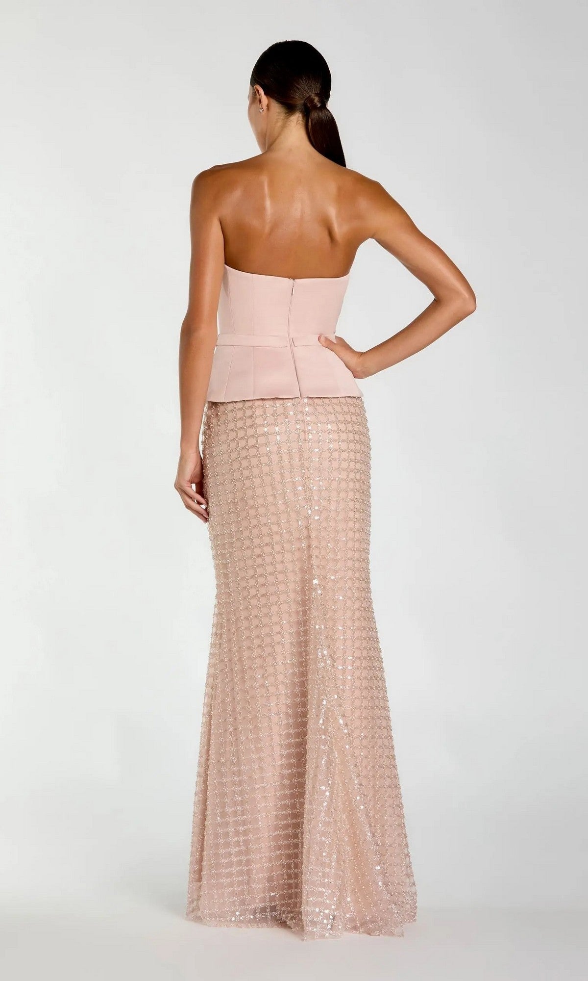 A woman stands with her back to the camera, wearing the Long Formal Dress: Mac Duggal 21227—a strapless, fitted blush pink gown featuring a peplum waist and a floor-length sequin skirt. Her hair is styled in a low ponytail.