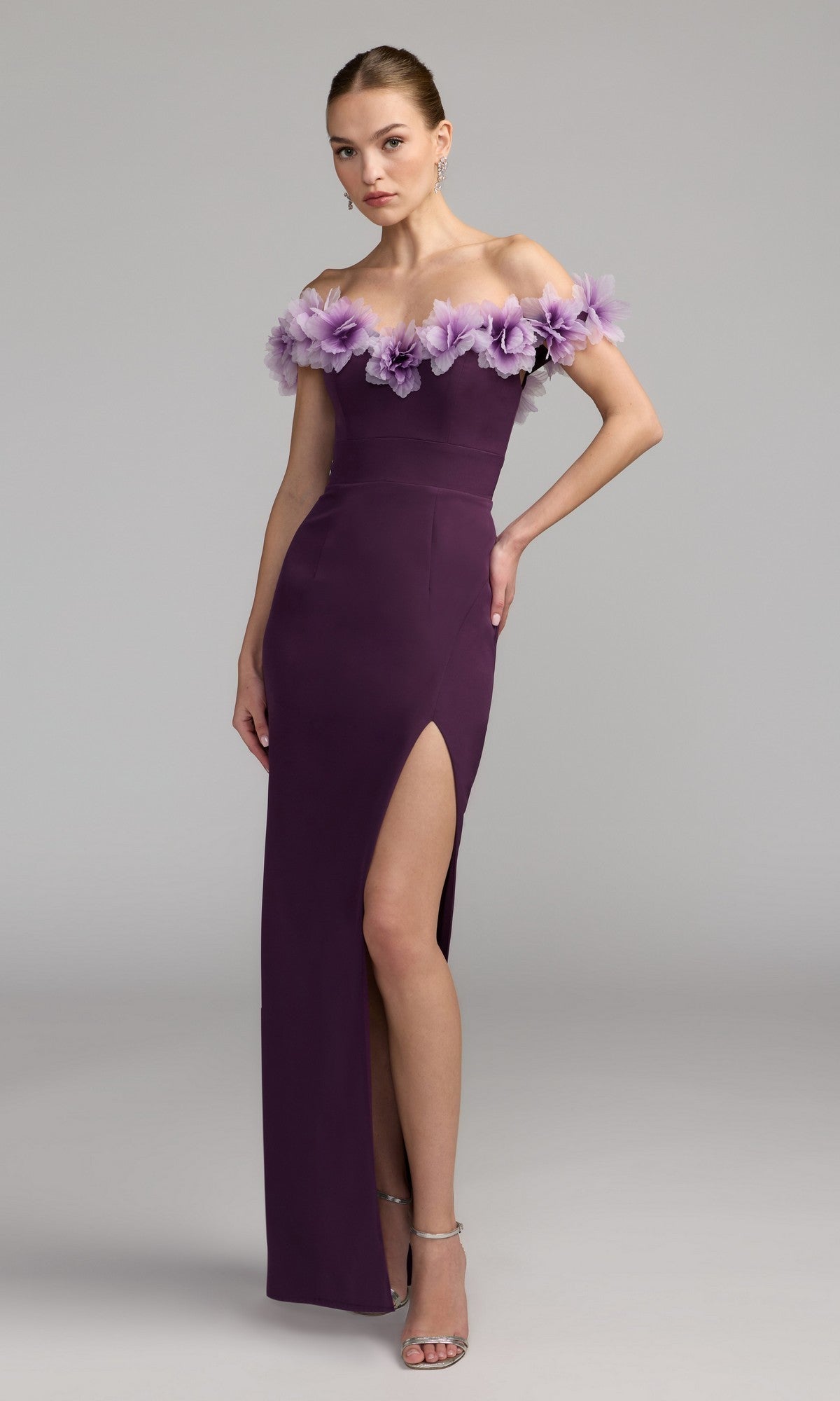 A woman in the Mac Duggal 21235 long formal dress in plum, featuring floral details and a thigh-high slit, with an elegant off-the-shoulder sweetheart neckline, posing against a plain gray background.