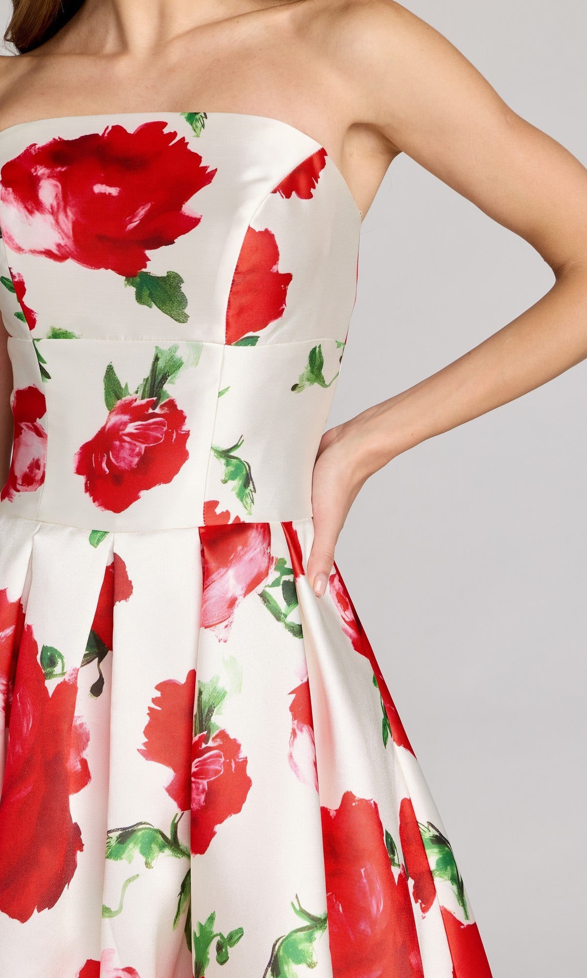 A woman poses with one hand on her hip against a neutral background, showcasing the Mac Duggal 21238 Tea-Length Party Dress—a white strapless midi featuring a vibrant red and green rose print.