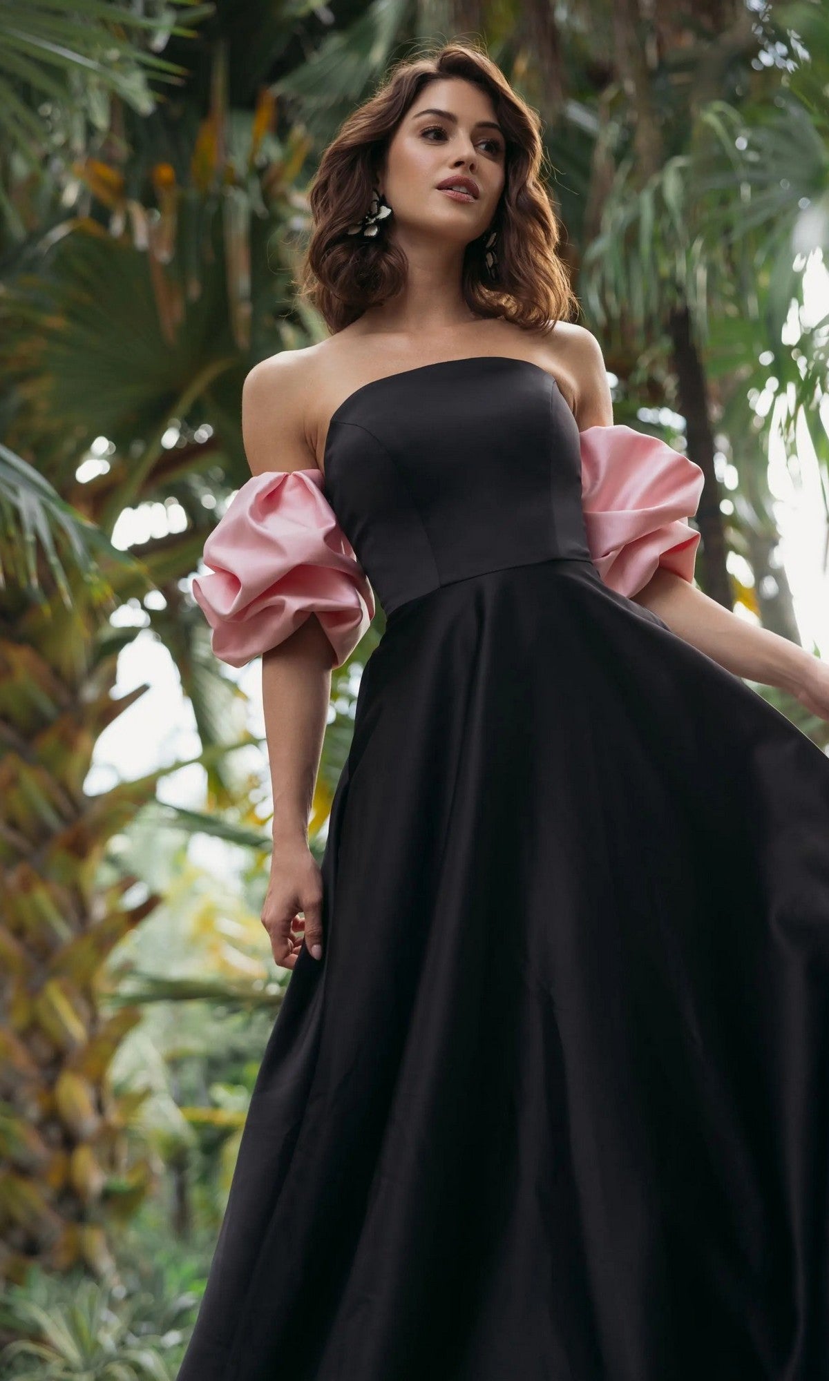 A woman stands outdoors in front of palm trees, wearing the Long Formal Dress: Mac Duggal 21241—a stunning black strapless satin gown featuring voluminous pink off-the-shoulder sleeves.