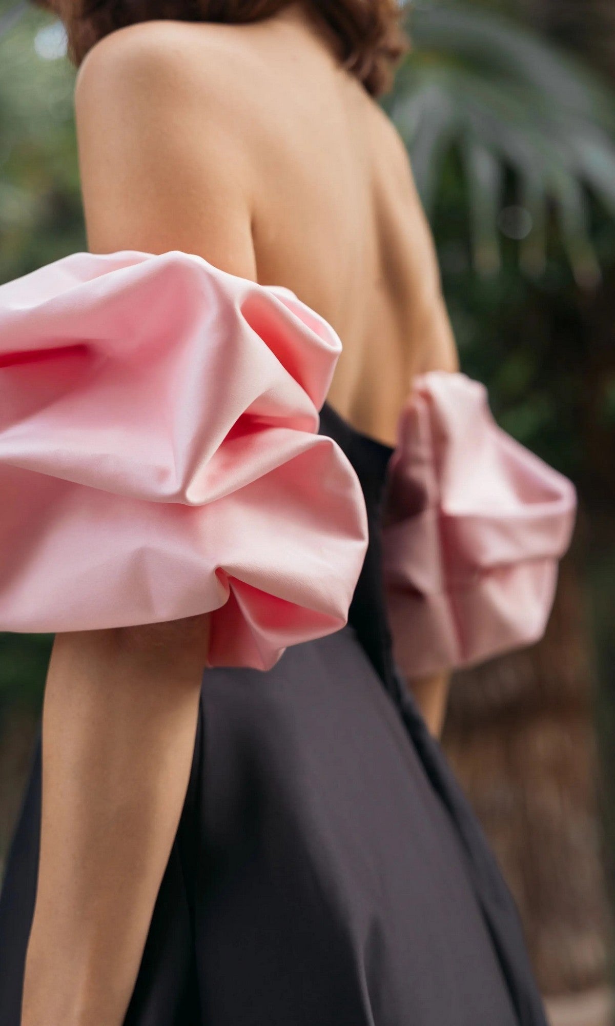 A woman stands outdoors with blurred greenery in the background, wearing the Long Formal Dress: Mac Duggal 21241—a black strapless satin gown featuring large pink satin puff sleeves.