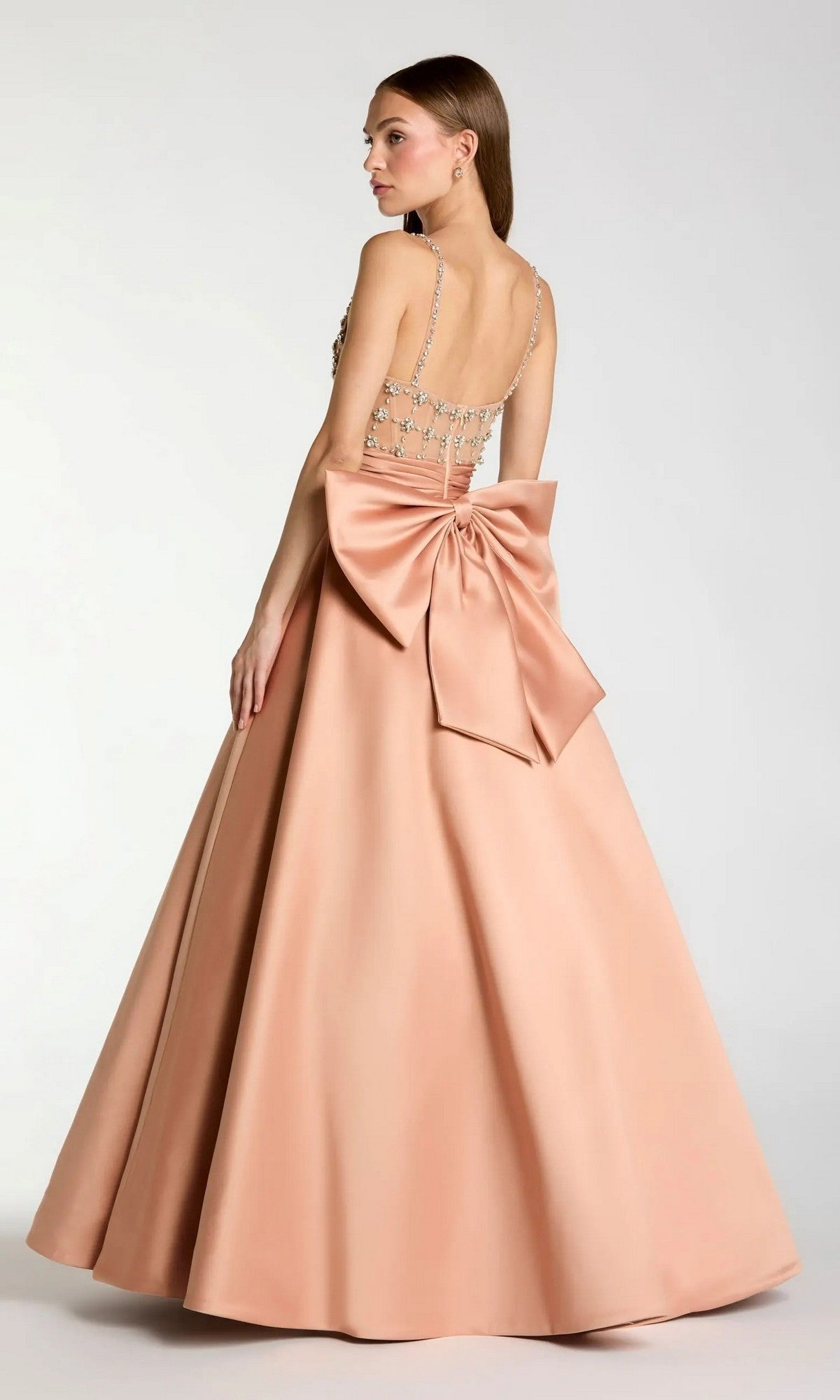 A woman is seen from behind wearing the Mac Duggal 21242 Long Formal Dress—a floor-length, peach satin ballgown featuring crystal straps and a dramatic bow detail on the back, set against a plain background.