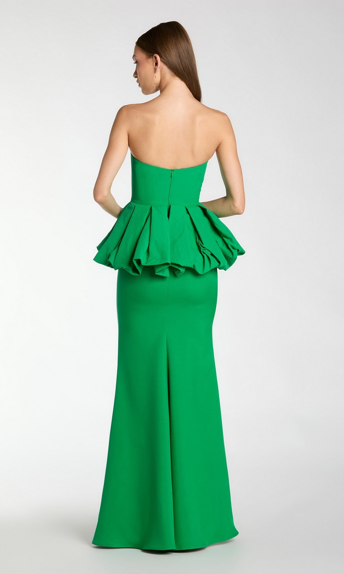 A woman stands facing away from the camera against a plain background, wearing the Mac Duggal 21243 Long Formal Dress—a green strapless gown with a ruffled peplum detail at the waist and a subtle trumpet silhouette.