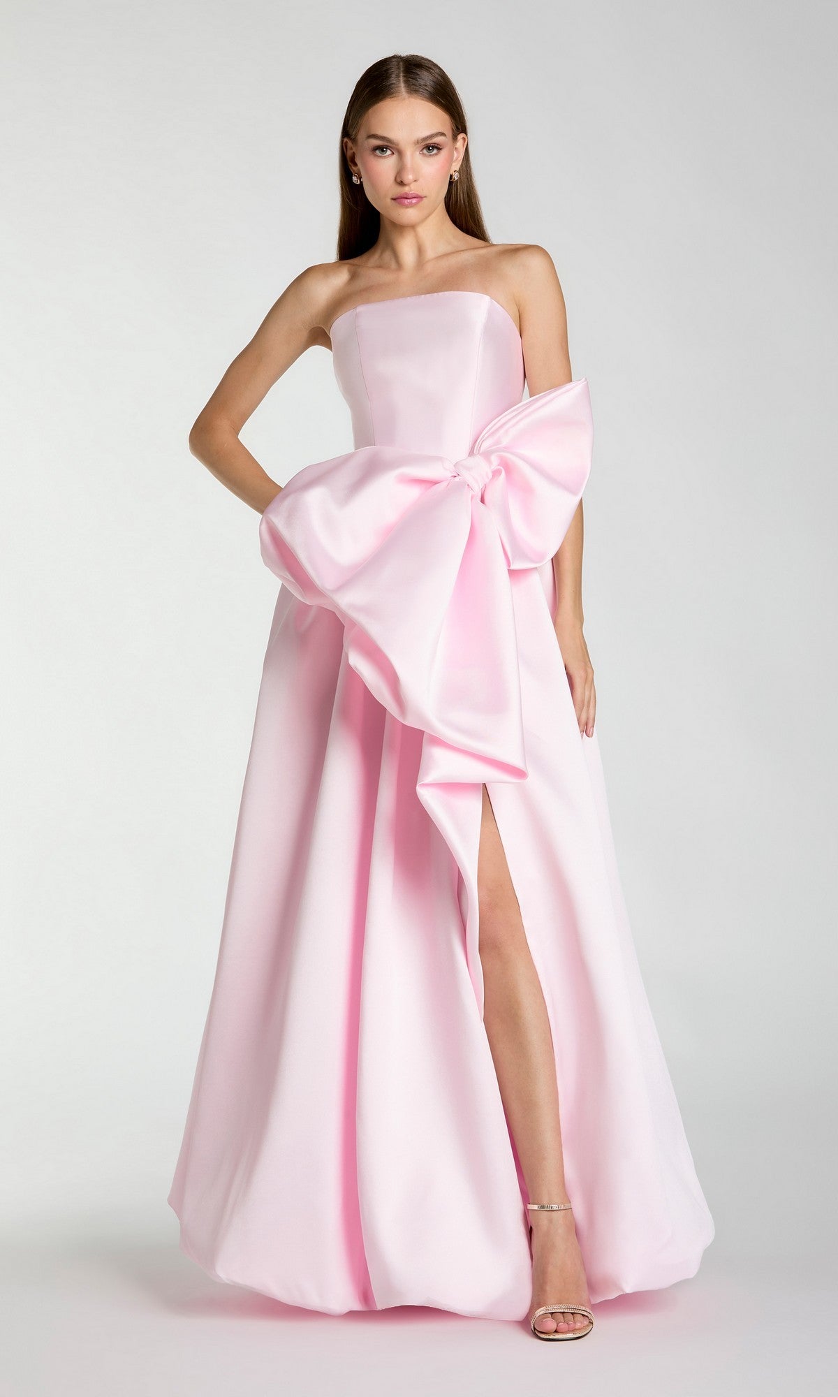 A woman wearing the Mac Duggal 21245 long formal dress in ice pink, featuring a strapless design, floor-length silhouette, large bow detail at the waist, and a thigh-high slit, stands against a plain background.