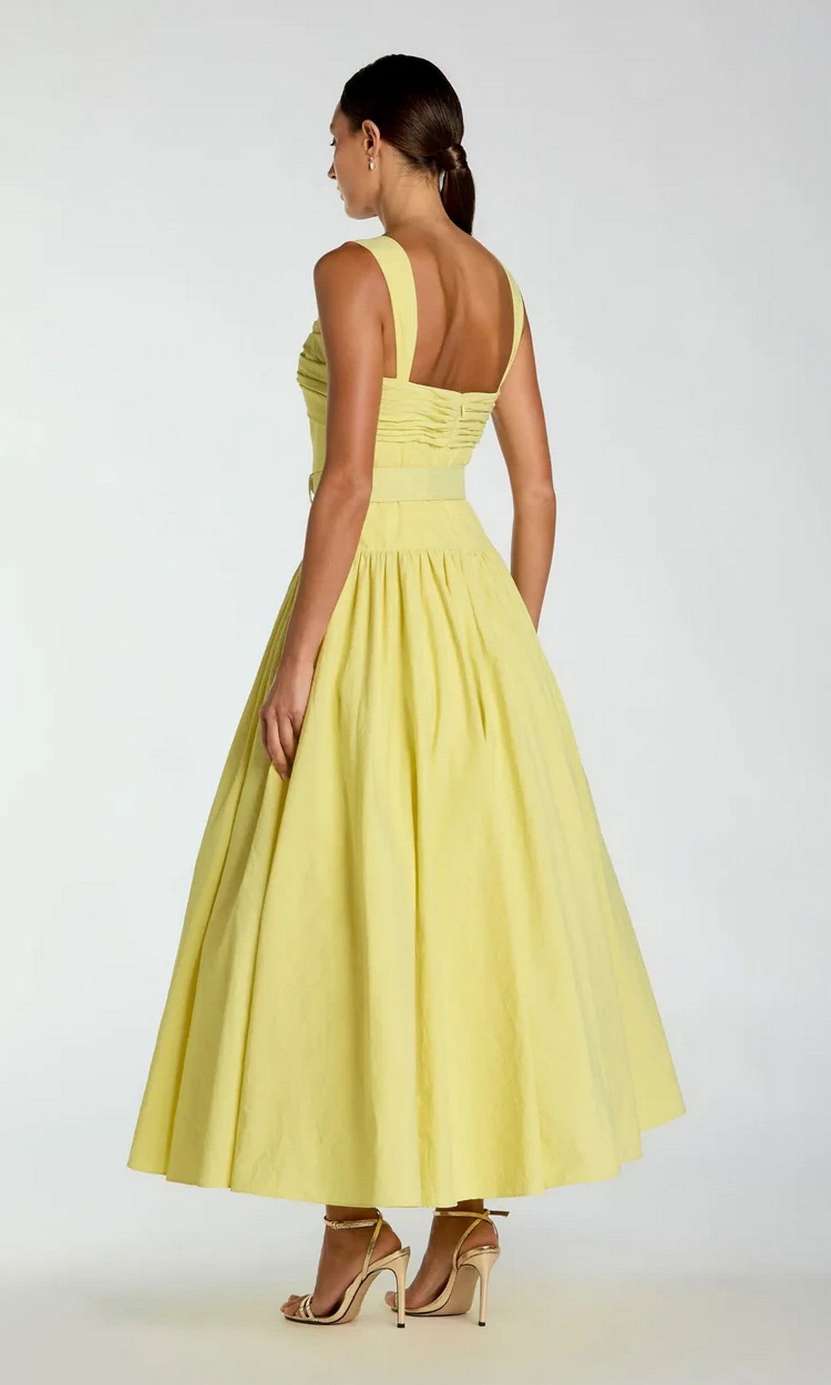 A woman stands in profile with her back to the camera, wearing the Tea-Length Party Dress: Mac Duggal 21251—a pale yellow, sleeveless, mid-length dress featuring a fitted bodice and flared skirt—paired with nude high-heeled sandals.