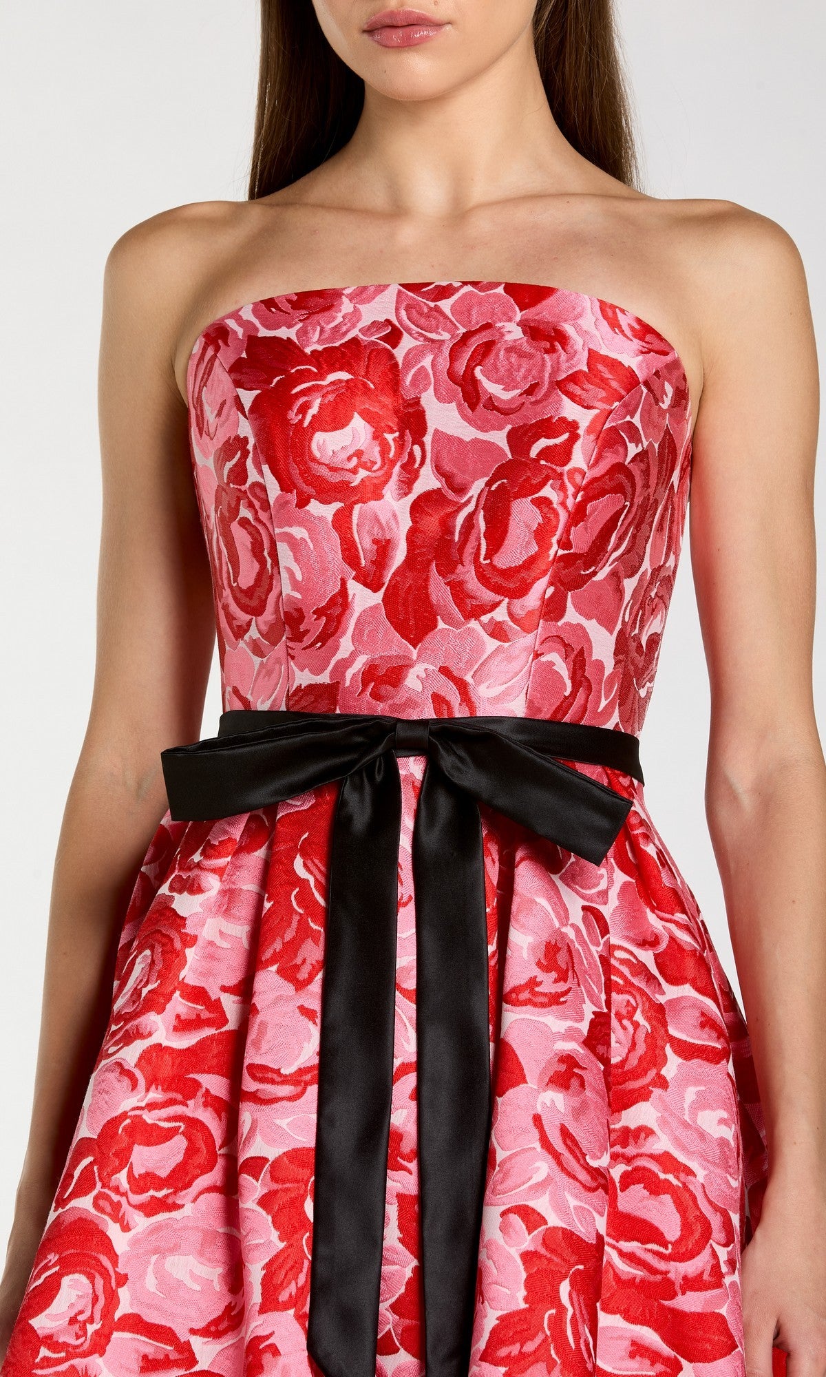 A woman is shown from shoulders to knees wearing the Tea-Length Party Dress: Mac Duggal 21252—a strapless pink dress featuring a bold red floral pattern and a black ribbon cinched at the waist.