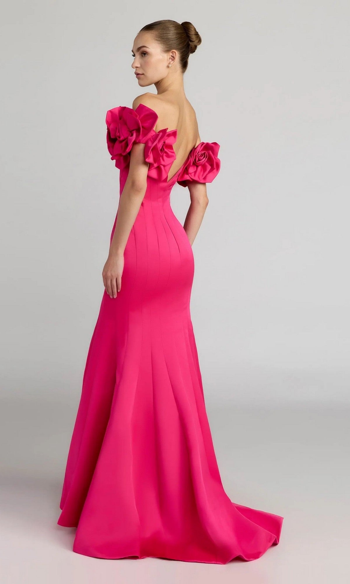 A woman stands against a plain background, looking over her shoulder in the sophisticated Mac Duggal 21263: a floor-length, hot-pink satin formal dress with off-the-shoulder ruffled sleeves—an ensemble that exudes elegance.