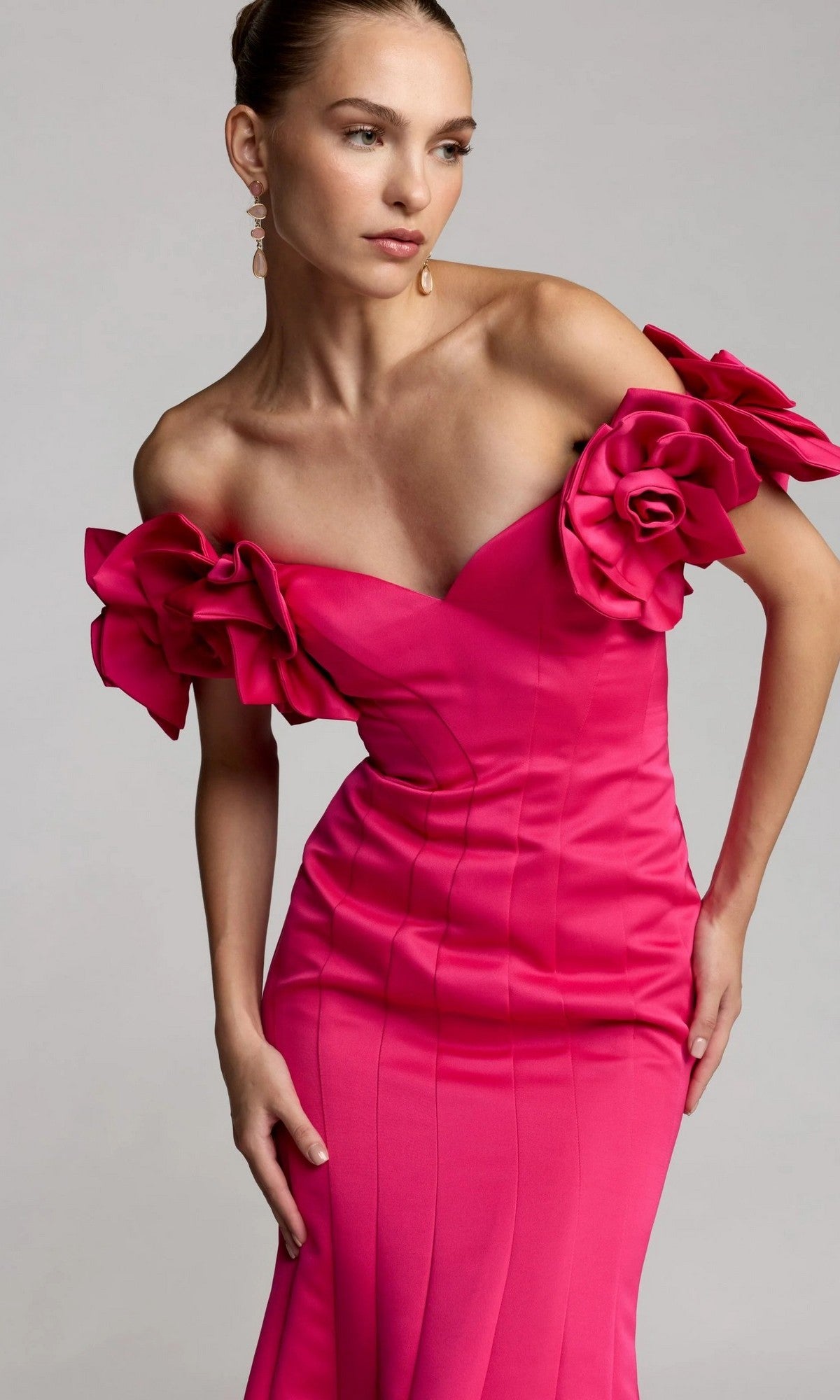 A woman models the Mac Duggal 21263 long formal dress, featuring an off-the-shoulder neckline in hot pink satin with dramatic floral accents on the sleeves, standing against a plain background.