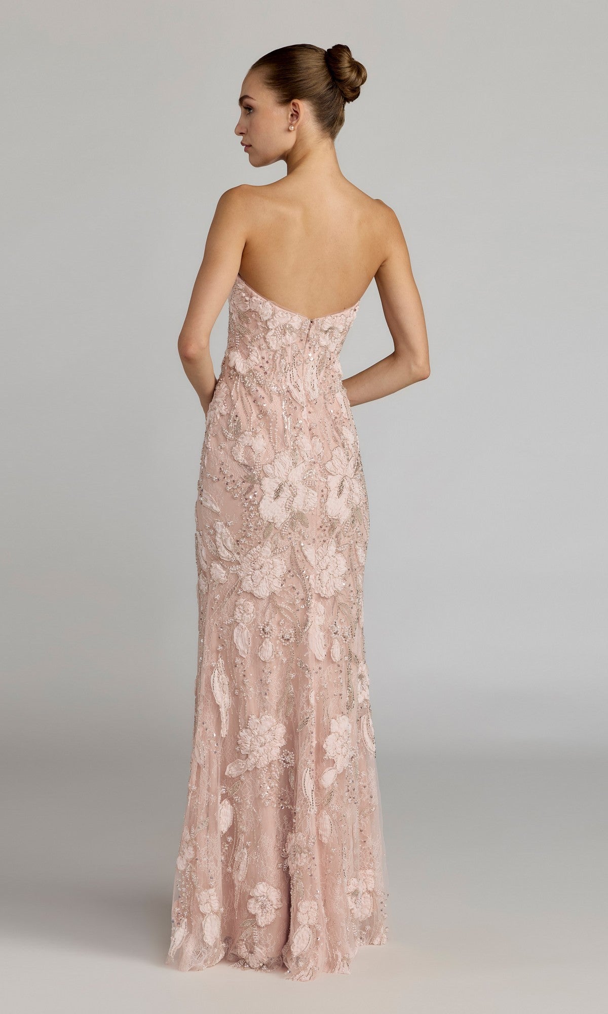 A woman stands with her back to the camera against a plain background, wearing the Mac Duggal 21283 long formal dress—a strapless, floor-length gown in light pink embellished lace.