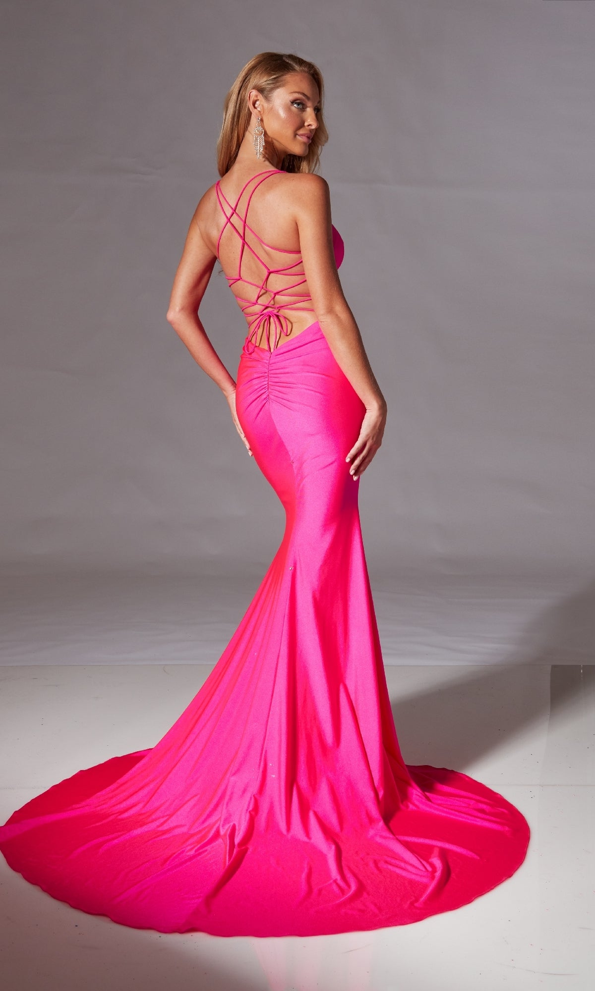 A woman models the Strappy Long Mermaid Prom Dress: Aerie Couture 212—a bright pink, floor-length gown with a lace-up open back and train—standing against a plain background.