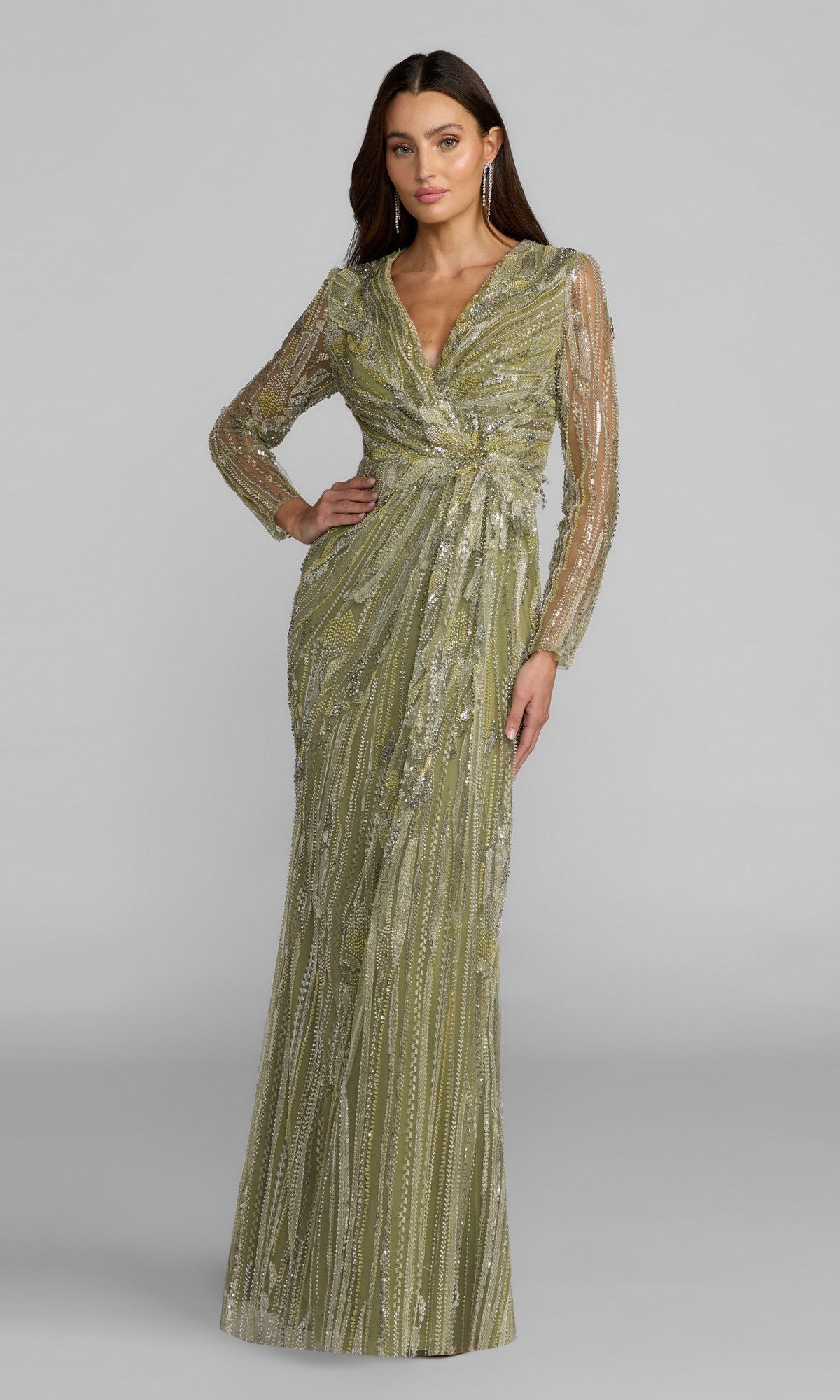 A woman stands wearing the Mac Duggal 21306 long formal dress in pistachio, featuring hand-embellished details, long sheer sleeves, and a wrap-style waist, posing against a plain gray background.