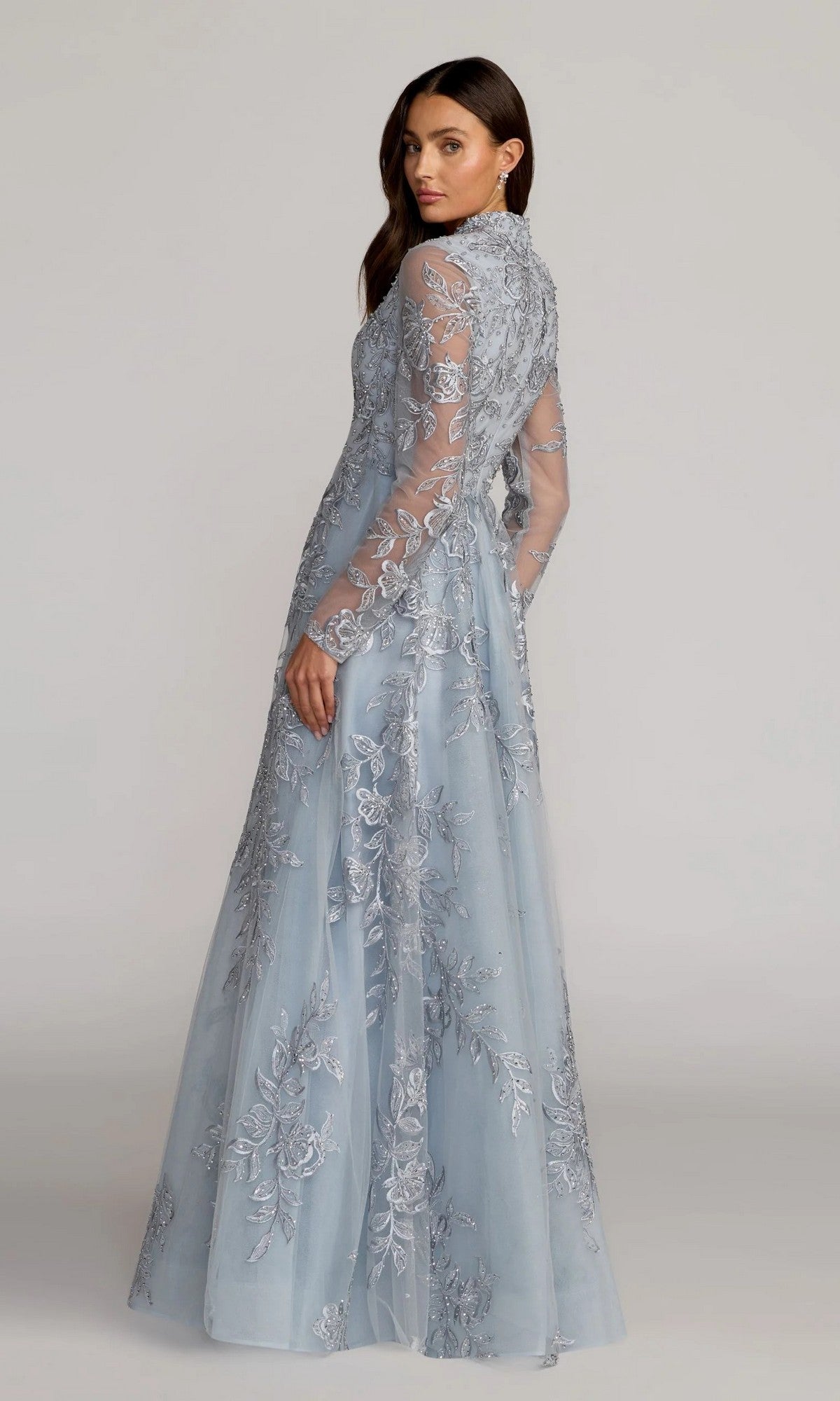Wearing the Mac Duggal 21307, a long formal dress in light blue with sheer long sleeves and intricate floral-beaded embroidery, a woman stands looking over her shoulder.