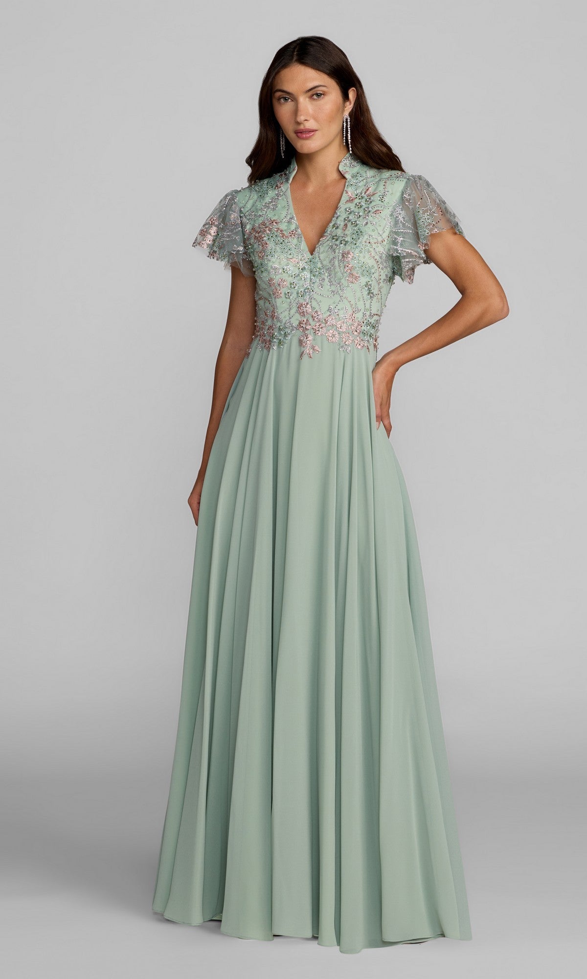 A woman wearing the Mac Duggal 21314 long formal dress in sage, featuring a floor-length silhouette, lace bodice, short flutter sleeves, V-neckline, and delicate floral detailing.