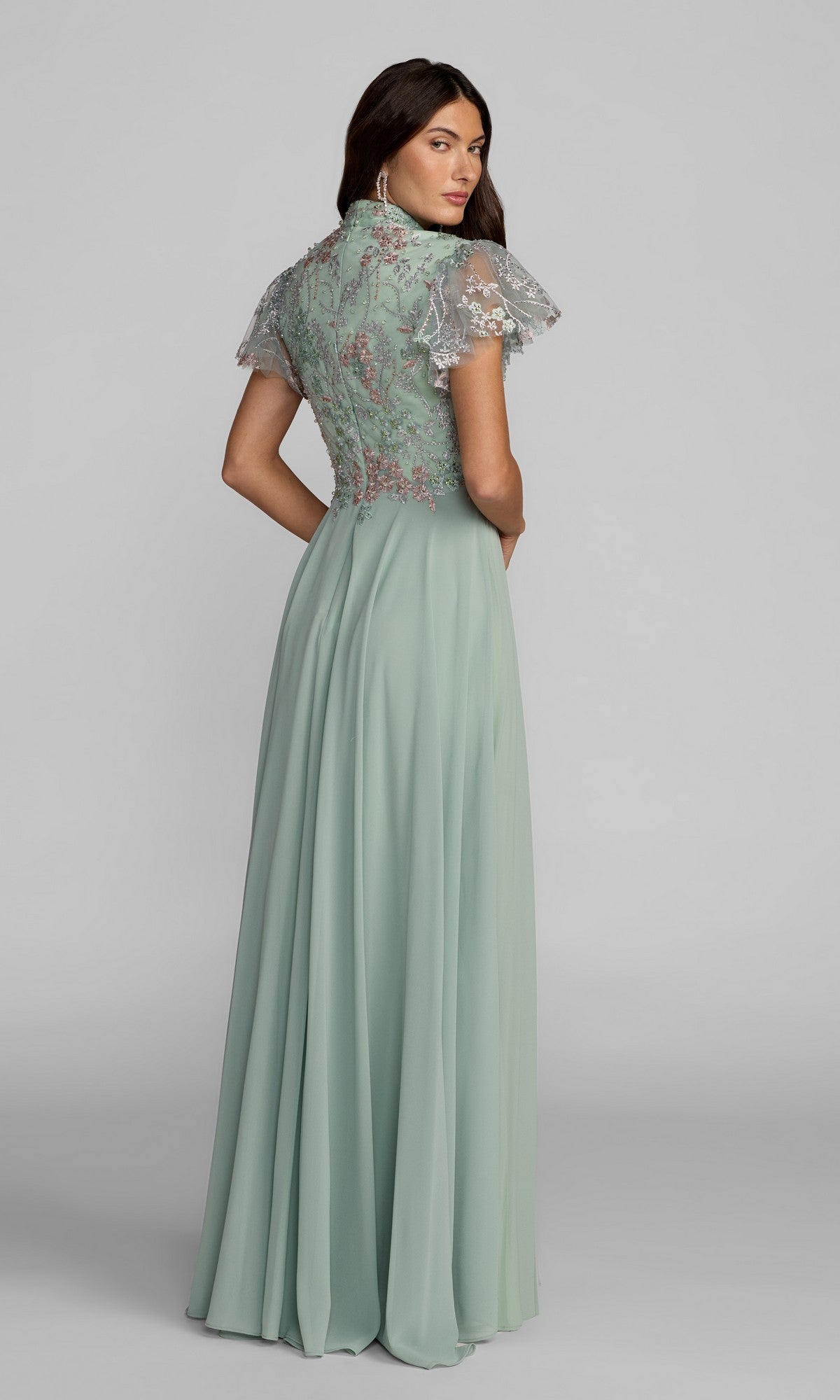 A woman stands looking over her shoulder in the Mac Duggal 21314 long formal dress—featuring a sage green hue, floral embroidery on the bodice, and short sheer sleeves.