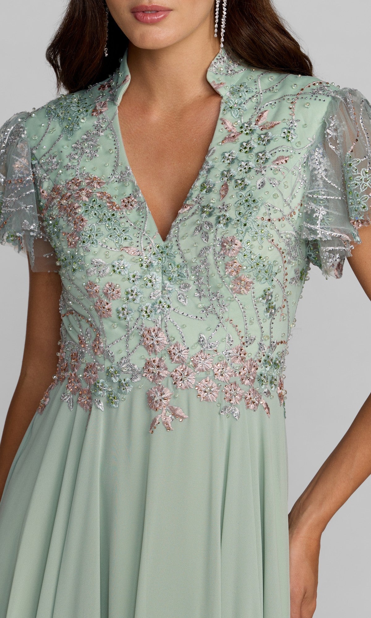 A woman models the Long Formal Dress: Mac Duggal 21314 in mint green, featuring short sheer sleeves, a high V-neckline, and silver and pink floral embroidery, with her hand resting on her hip.