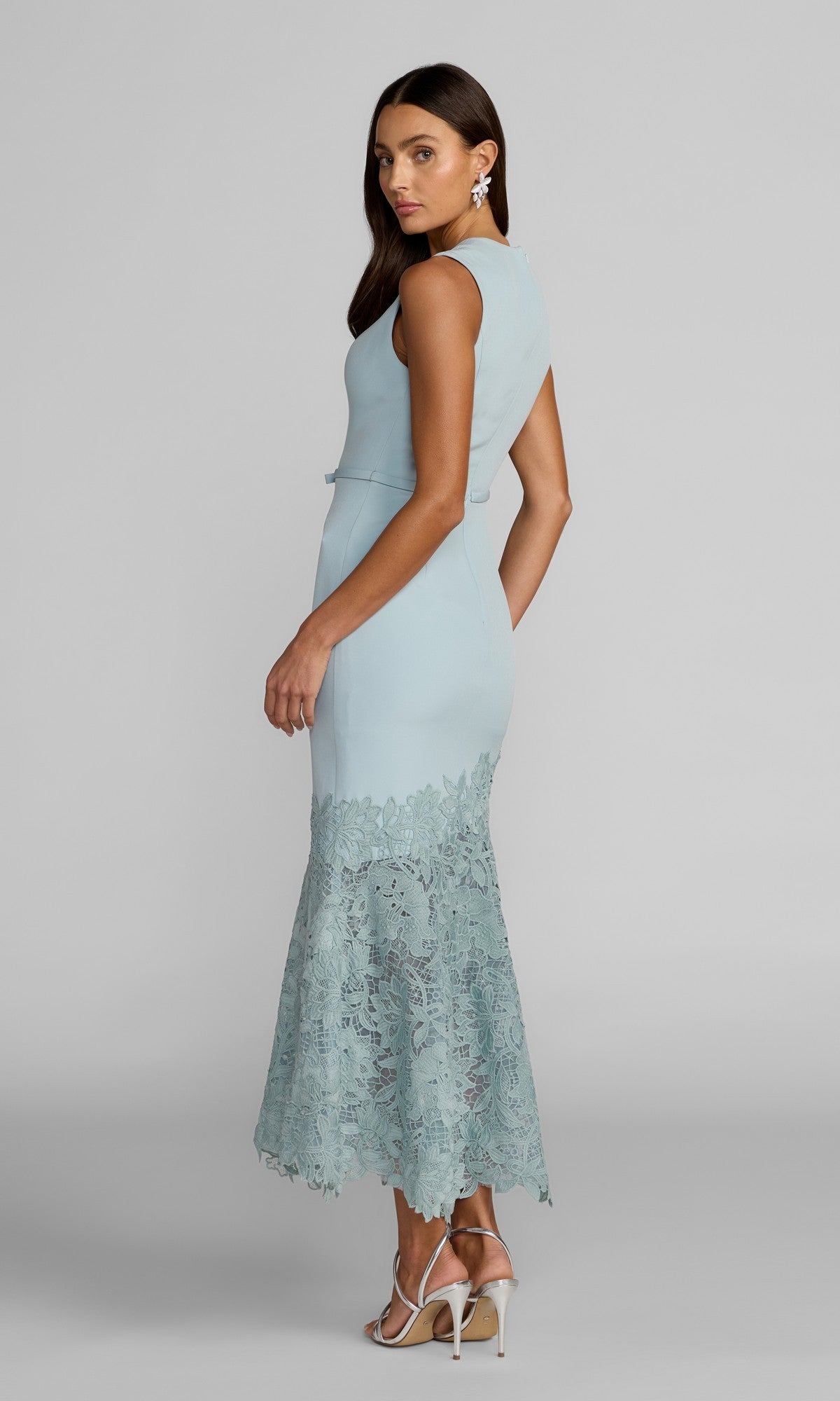 A woman stands sideways and looks over her shoulder, wearing the Mac Duggal 21355 Tea-Length Party Dress—a sleeveless crepe dress with lace detailing at the hem and an asymmetrical lace skirt—styled with white heels and statement earrings.