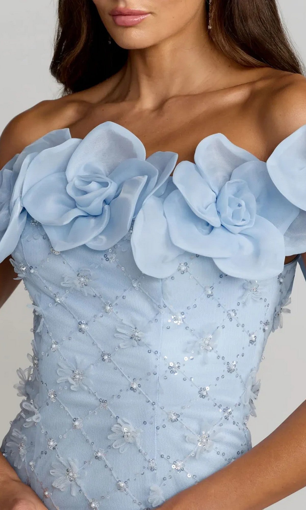 A woman is wearing the Mac Duggal 21363 midi party dress, featuring an off-the-shoulder design in light blue with large fabric flowers and sequin-lattice motifs embellishing the bodice.