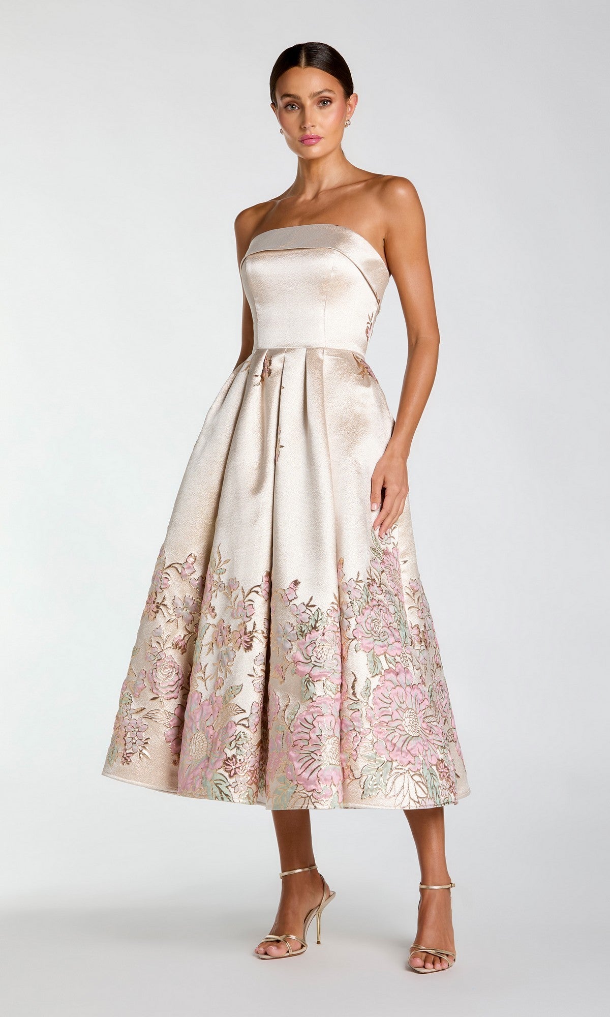 A woman is wearing the Mac Duggal 21372 tea-length party dress in Champagne Multi, featuring a strapless design and pink floral embroidery, paired with high-heeled sandals and standing against a plain light background.