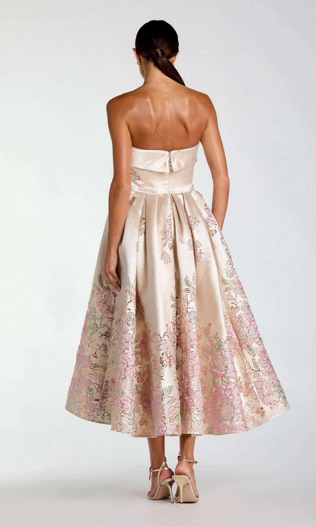 A woman stands with her back to the camera wearing the Tea-Length Party Dress: Mac Duggal 21372—a gorgeous strapless brocade midi in light pink and gold floral tones—paired with matching high-heeled sandals.