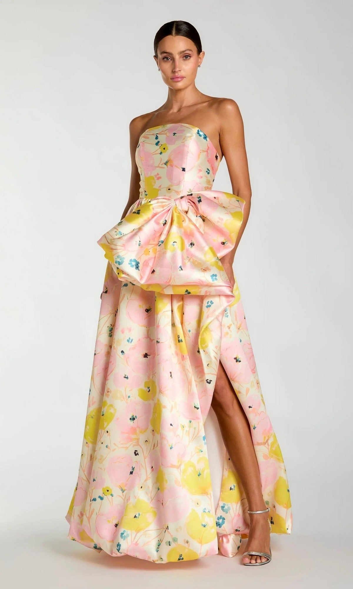 A woman wearing the Mac Duggal 21376 long formal dress—a strapless Mikado ballgown adorned with a pastel floral pattern, featuring a peplum waist detail, an oversized bow accent at the waist, and a dramatic thigh-high slit—paired with strappy heels.