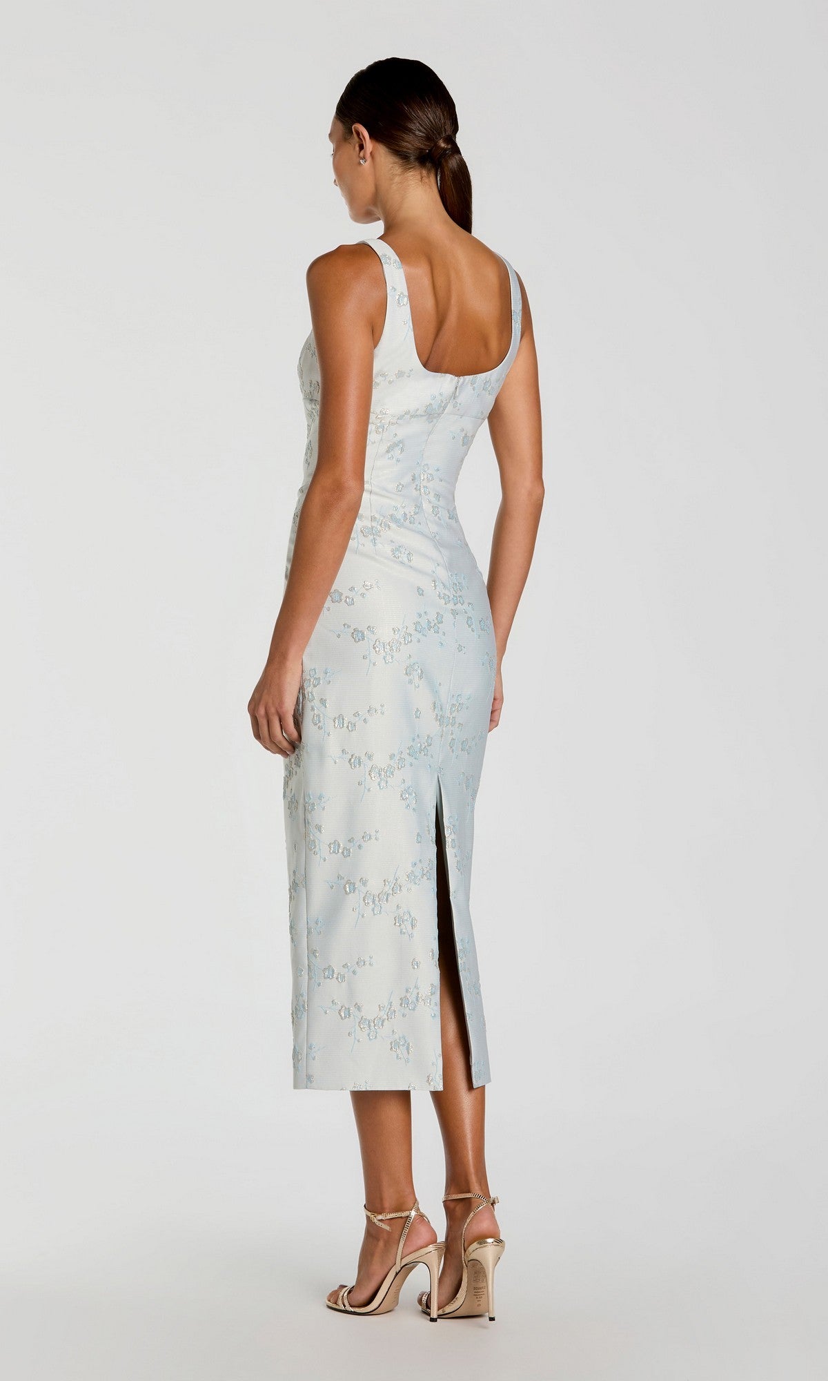 A woman is standing with her back to the camera against a plain background, wearing the Mac Duggal 21377 Midi Party Dress in light blue brocade featuring a back slit, paired with high-heeled sandals.