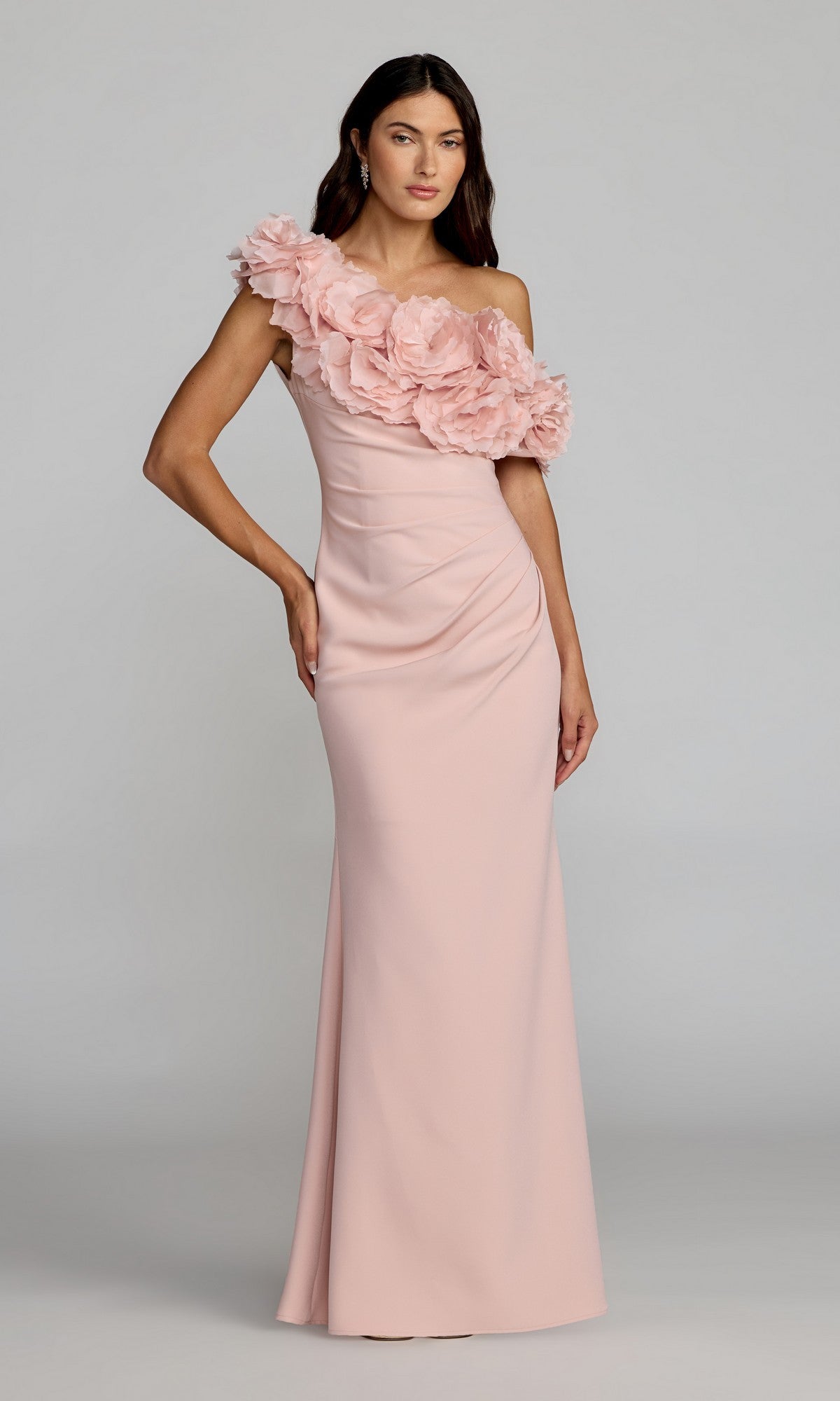 A woman wearing the Mac Duggal 21383 long formal dress in blush, featuring a floor-length one-shoulder design with dramatic 3D floral appliques cascading across the bodice, stands against a plain background.