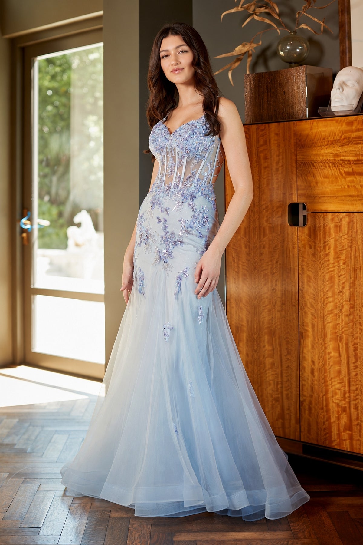 A model wears the Amelia Couture 2164, a floor-length, baby blue mermaid prom dress with a sheer, corset-style bodice and sequin flower appliques.