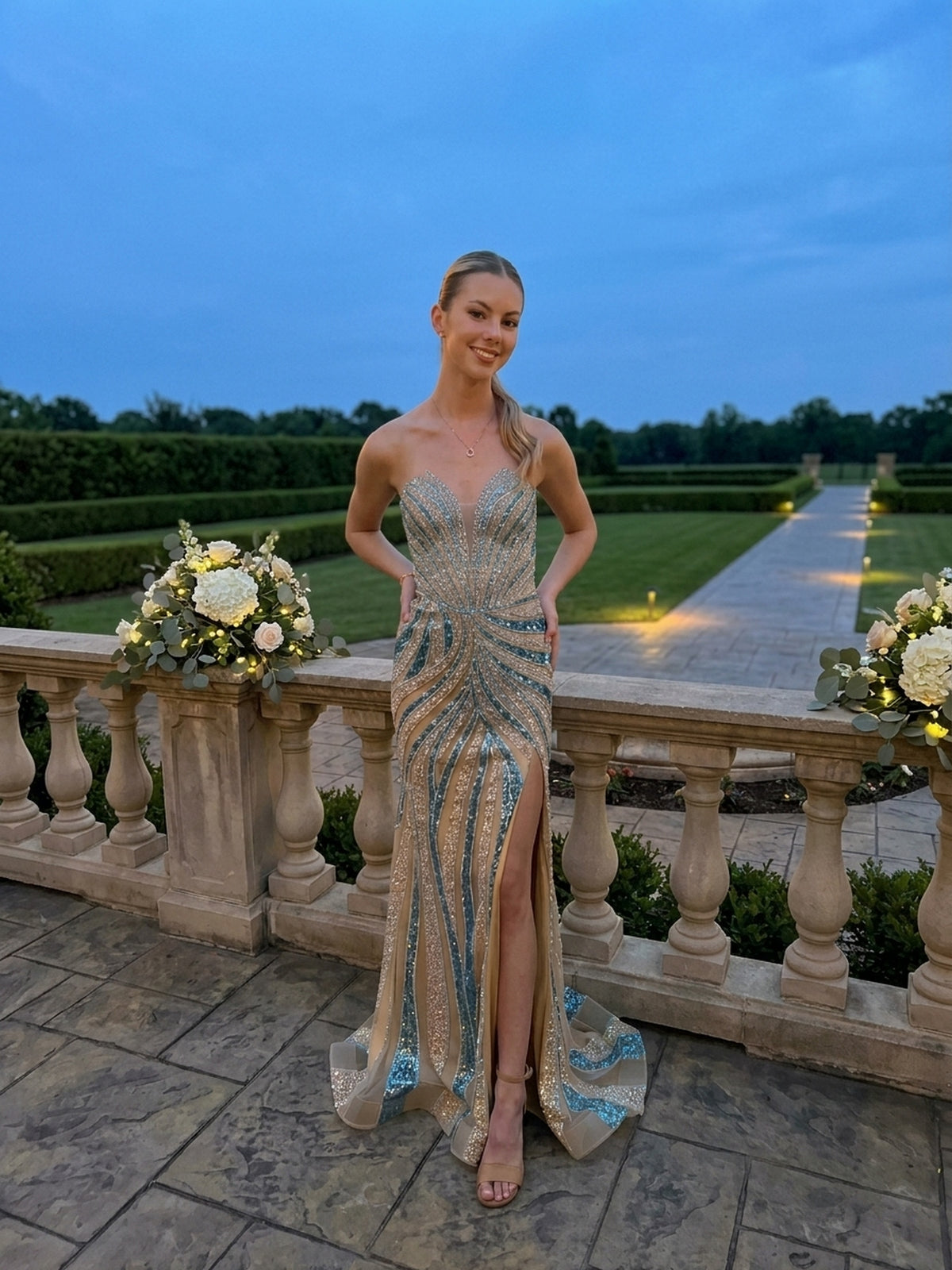 A person wearing the Long Prom Dress: Amelia Couture 2167, a retro glam, strapless, champagne and teal sequined floor-length gown with a thigh-high slit, stands on a stone terrace.