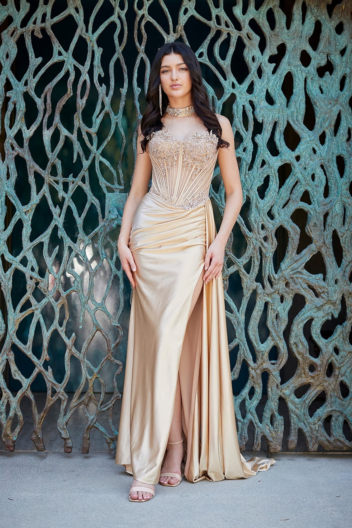 A woman wears a champagne-colored Long Prom Dress: Amelia Couture 2169, featuring a strapless beaded corset bodice and a high leg slit.