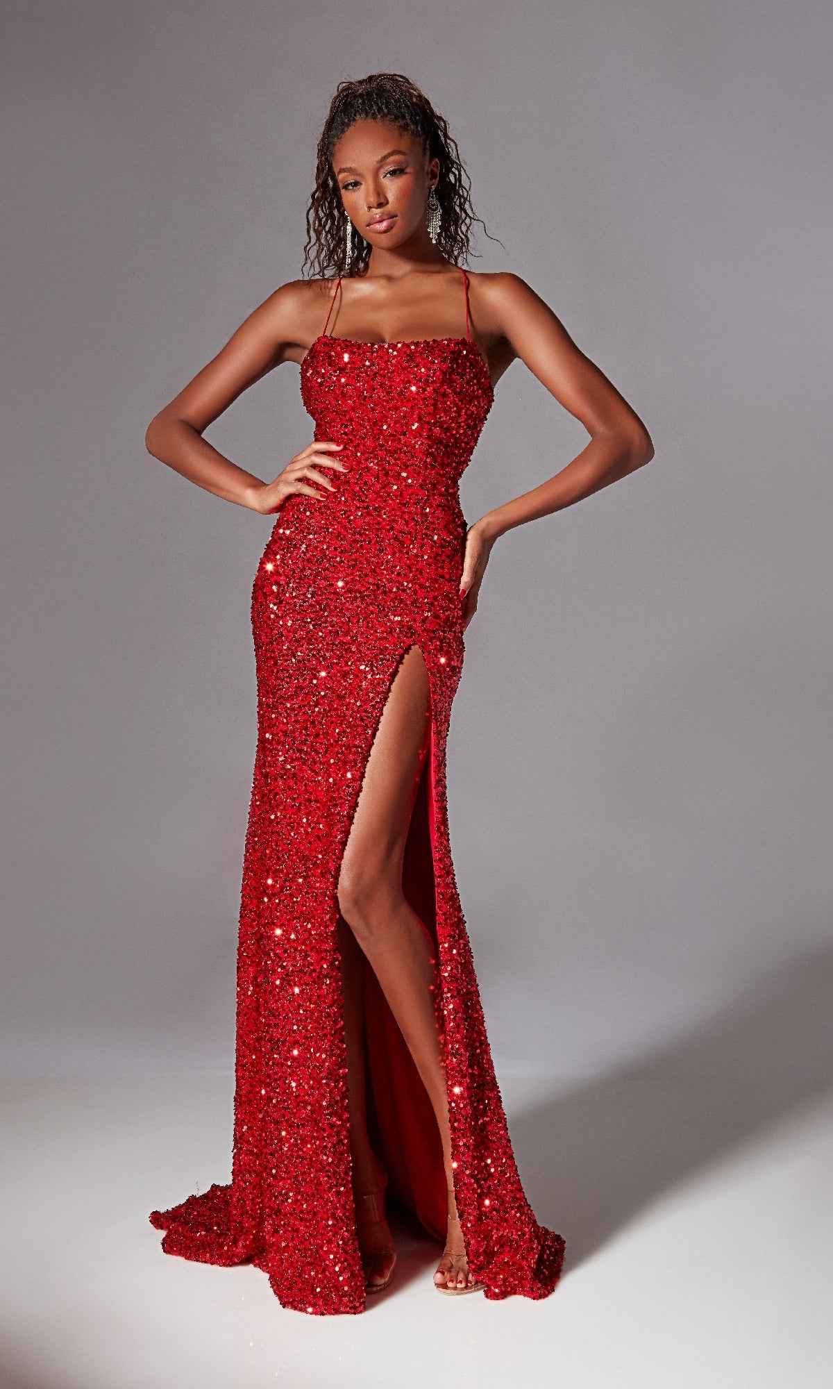 A woman models the Backless Long Sequin Prom Dress: Aerie Couture 217, featuring a fitted silhouette, thin straps, striking red sequins, and a thigh-high slit against a plain gray background.